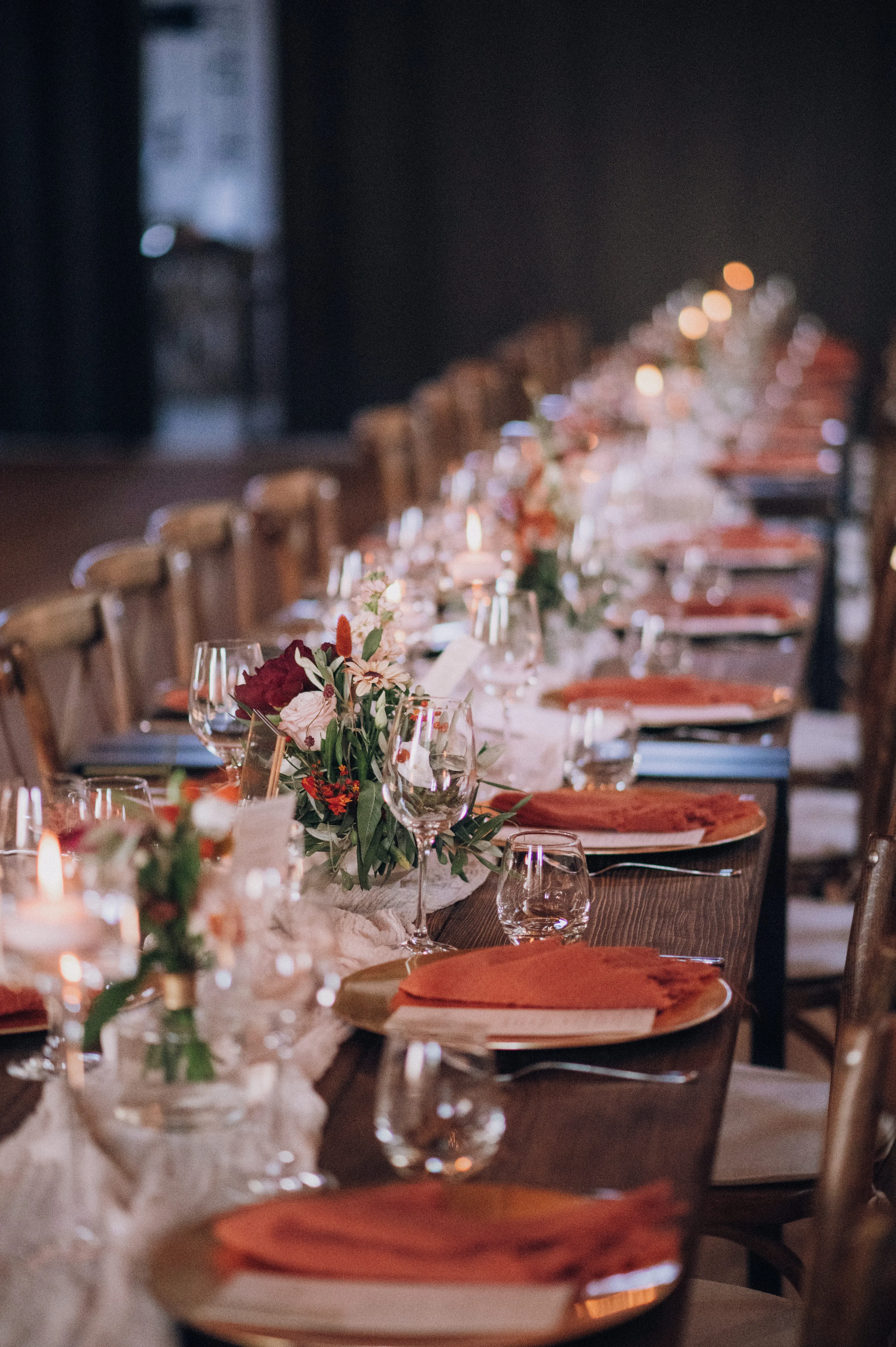 Ein langer Holztisch mit eleganten Gläsern, Tellern und orangefarbenen Servietten ist für eine Hochzeit gedeckt. In der Mitte befinden sich dezente Blumengestecke, die durch sanftes Kerzenlicht beleuchtet werden.