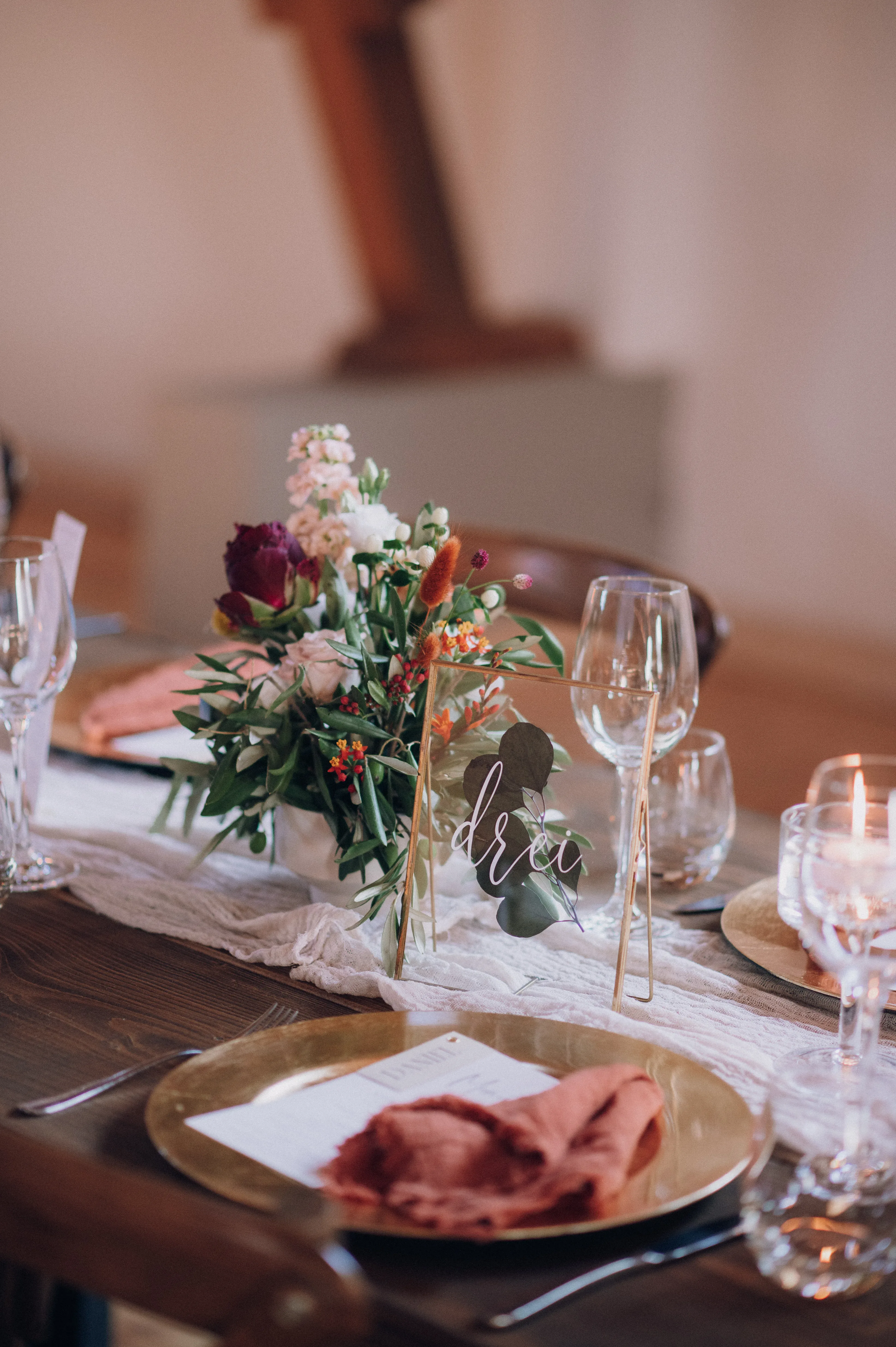 Nahaufnahme einer Hochzeitstafel, liebevoll dekoriert mit floralen Akzenten und einem Namensschild „Drei“. Goldene Teller und kunstvoll gefaltete Servietten in warmen Farben runden die elegante Tafel ab.
