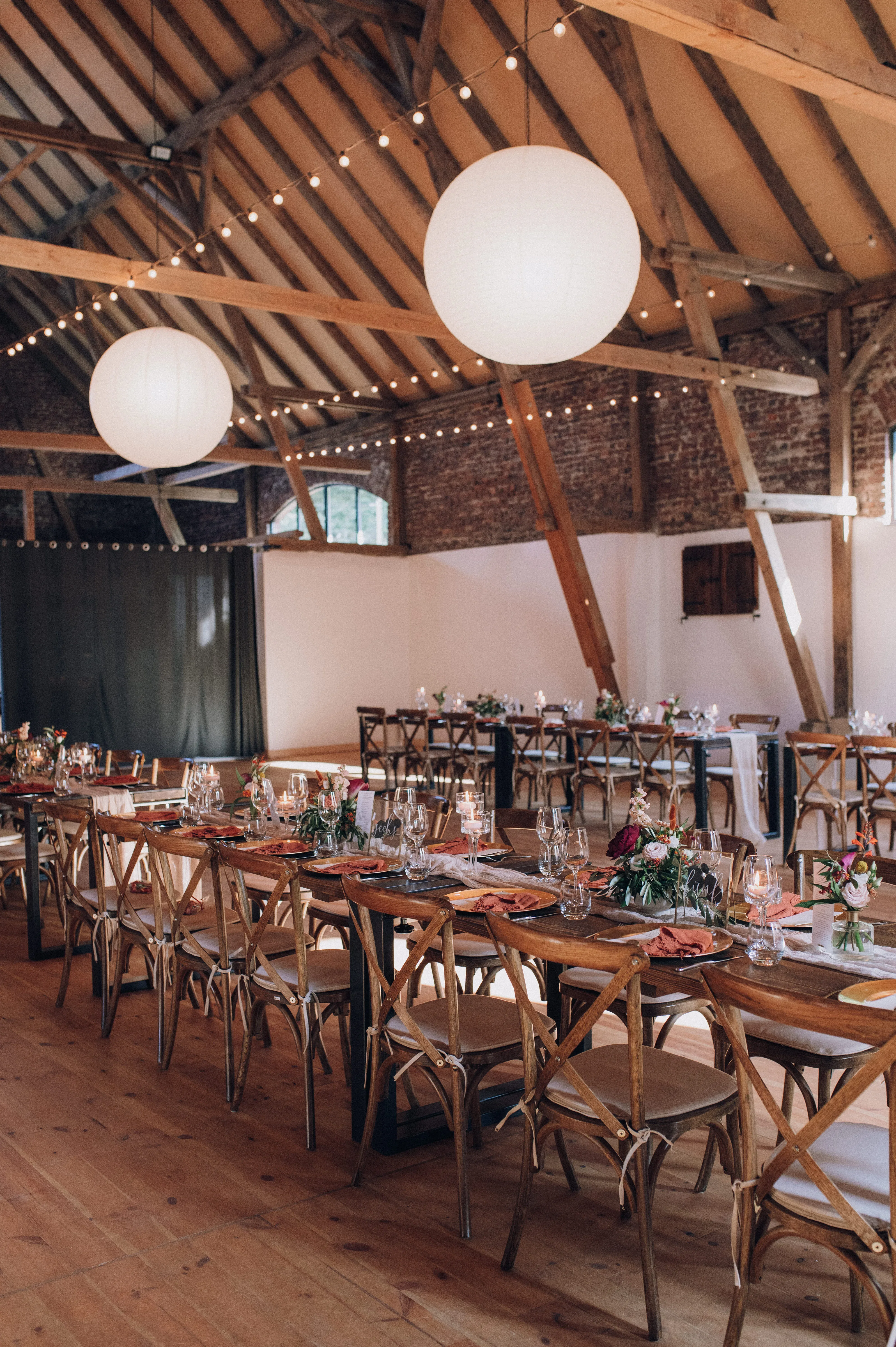 Hochzeitssaal im rustikalen Stil mit langen Tafeln, die für das Festessen gedeckt sind. Über den Tischen hängen weiße Papierlampions, die den Raum sanft erleuchten, während Kerzen auf den Tischen romantisches Licht spenden.