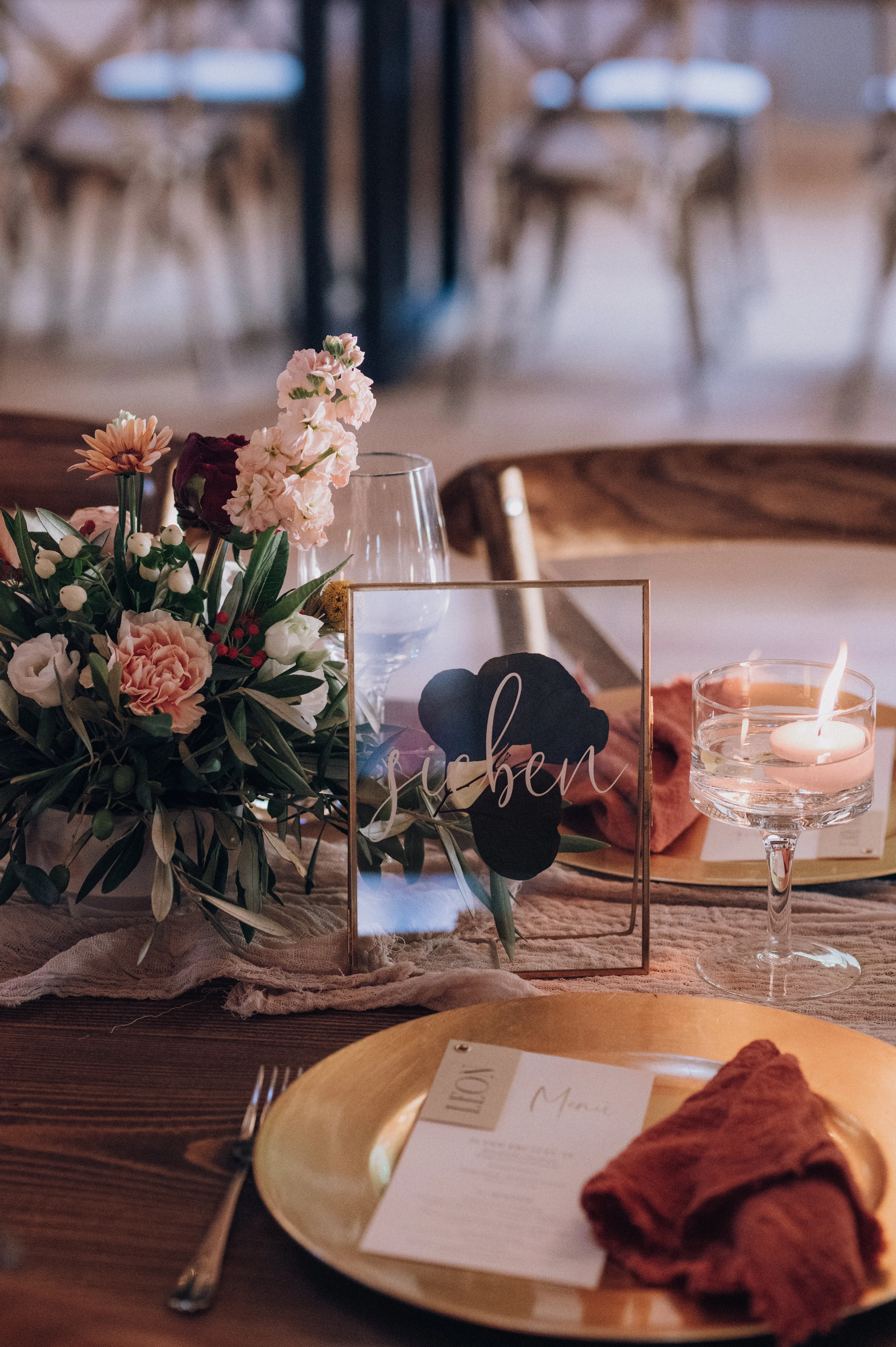 Ein liebevoll gedeckter Tisch mit goldfarbenem Geschirr, oranger Stoffserviette und einer brennenden Kerze im Glas. Ein kleines Blumengesteck in zarten Rosa- und Rottönen ziert die Mitte des Tisches.