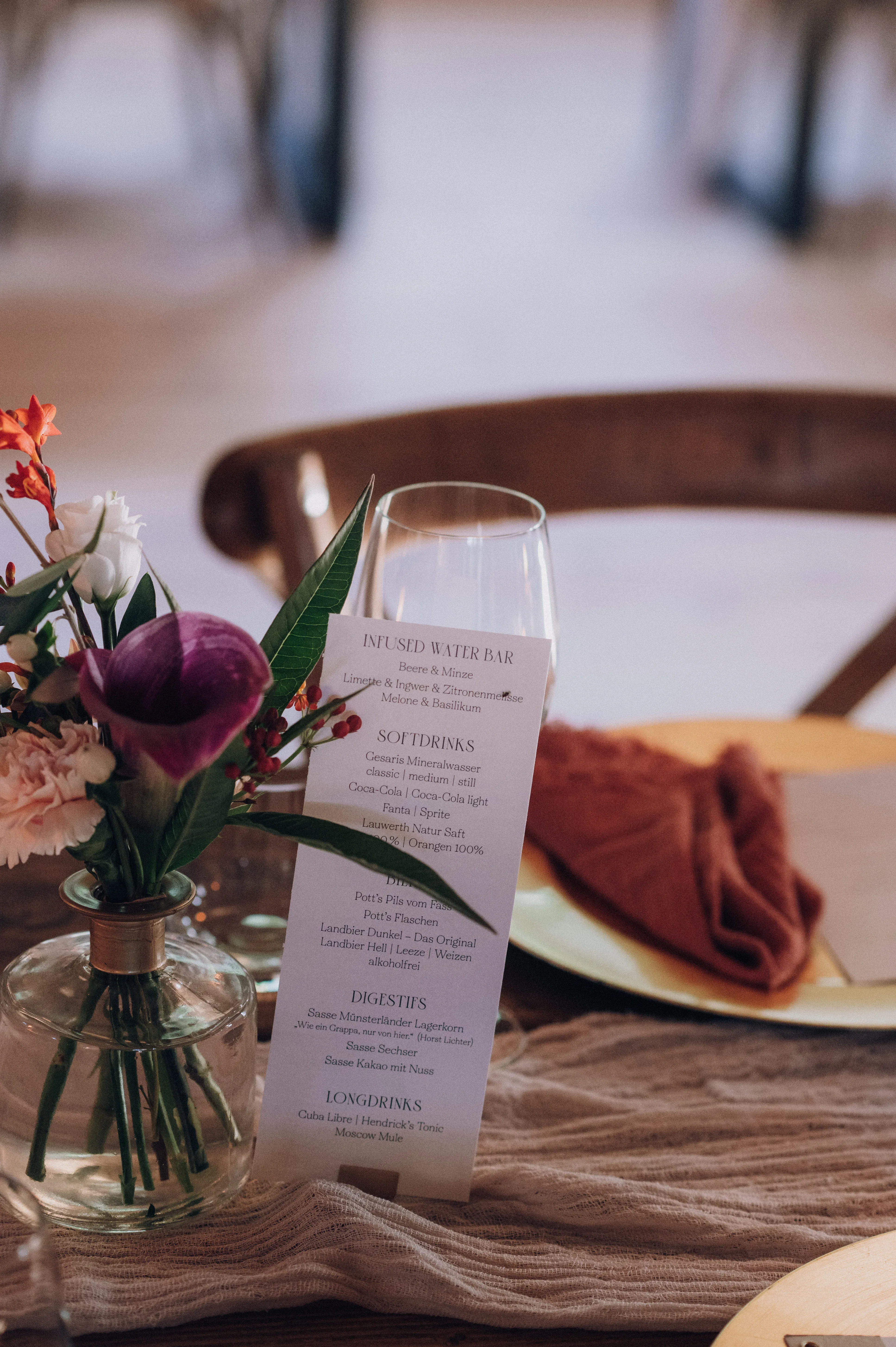 Eine Menükarte, umgeben von eleganten Weingläsern und Blumenarrangements, steht auf einem Hochzeitstisch mit rustikalem Holz und orangefarbenen Servietten.