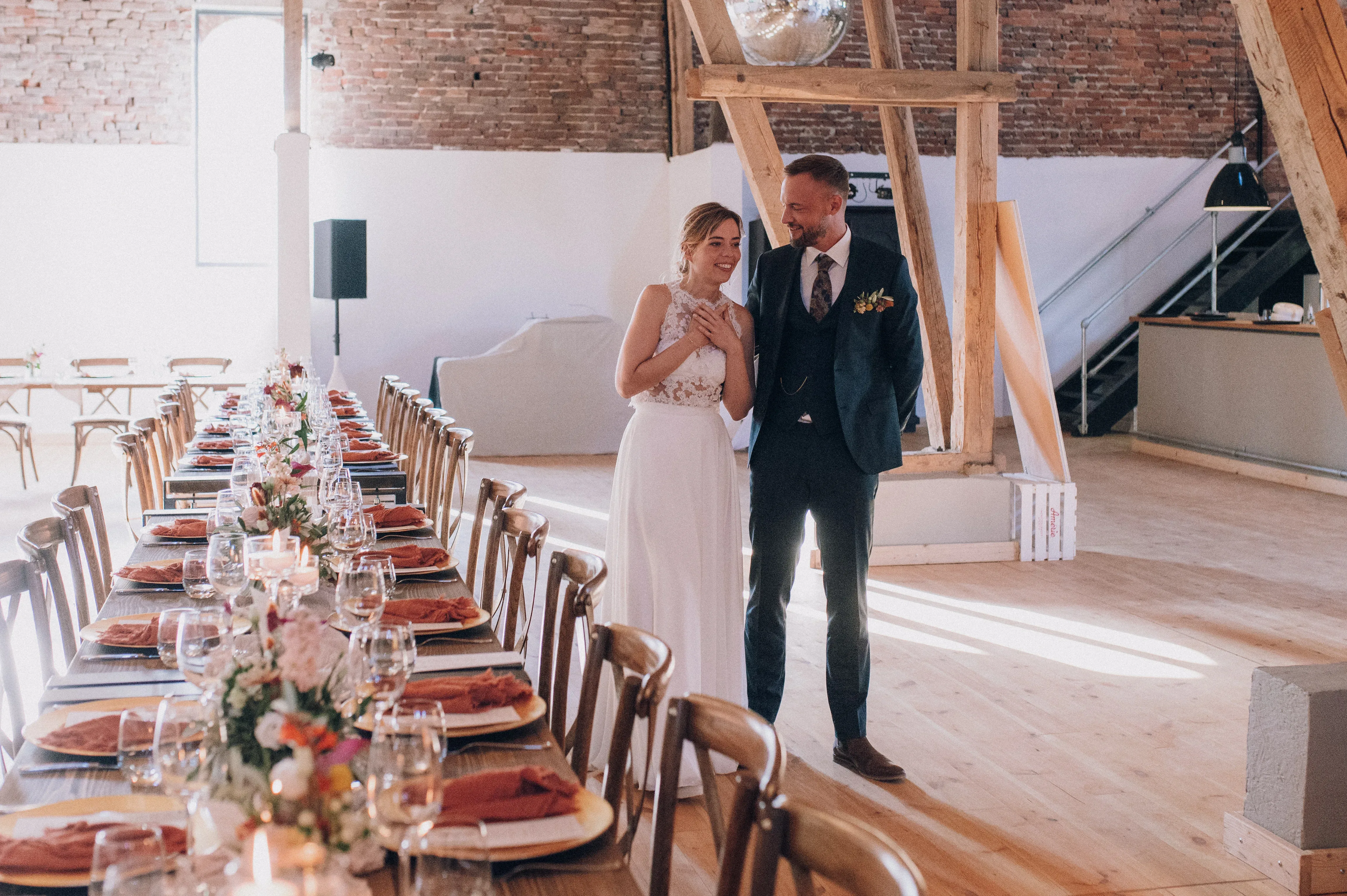 Das Hochzeitspaar betritt den festlich dekorierten Saal zum ersten Mal. Die Braut hält sich gerührt die Hand ans Herz, während der Bräutigam sie glücklich ansieht. Der lange, festlich gedeckte Tisch mit Kerzen und Blumenarrangements sorgt für eine warme Atmosphäre.