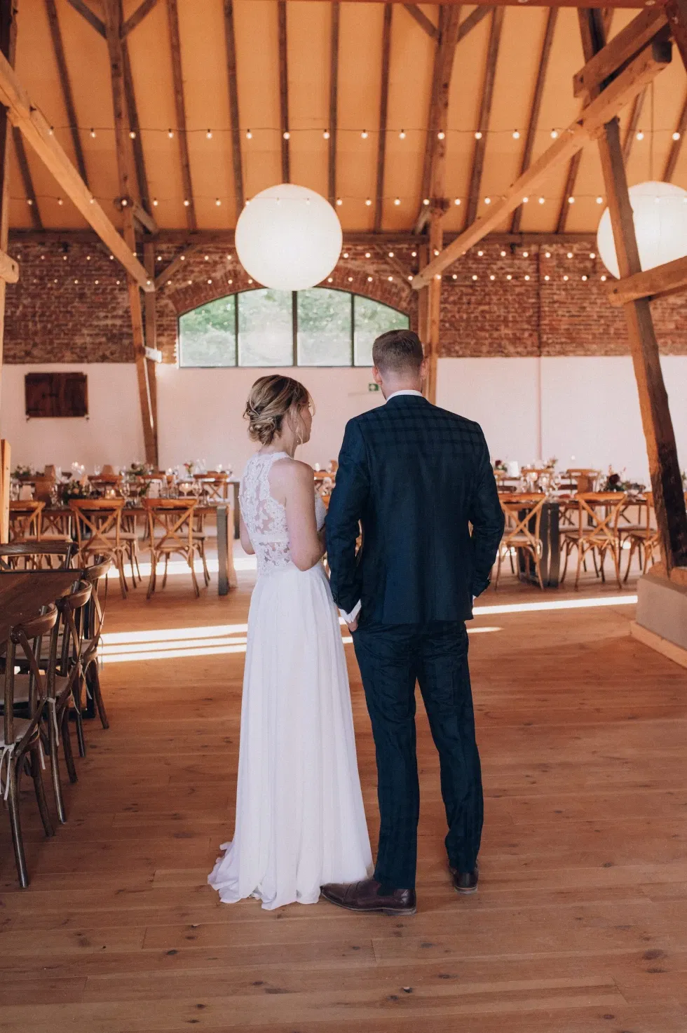 Ein Brautpaar steht Hand in Hand in einem stilvoll dekorierten Hochzeitssaal mit Holzbalken und runden Lampen.