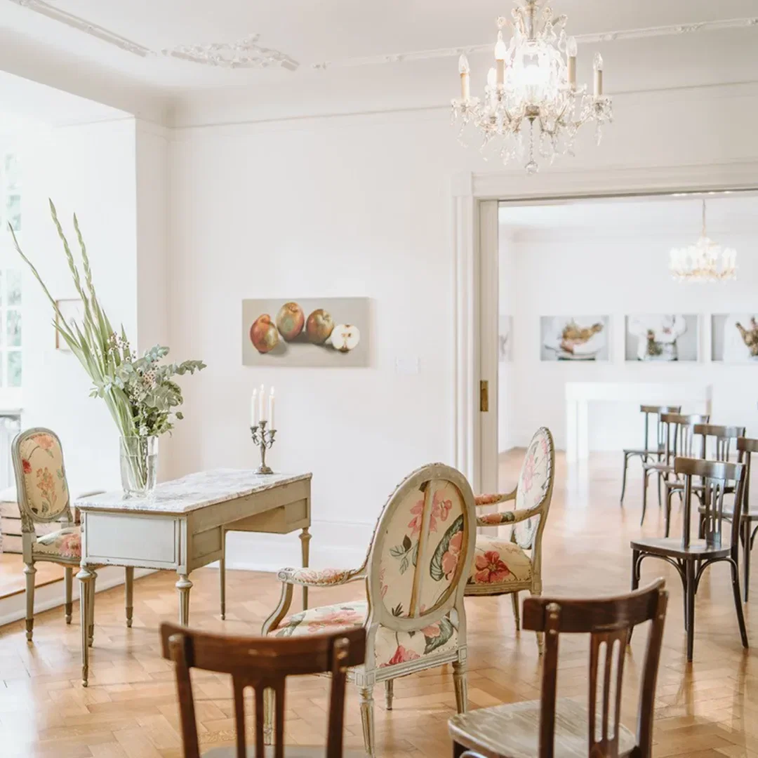 Innenansicht eines hellen und elegant eingerichteten Zimmers in Burg Lede, Bonn, mit weißen Wänden, Kronleuchtern, antiken Möbeln mit floralen Mustern und einem Schreibtisch mit Kerzenhalter und Blumenarrangement.