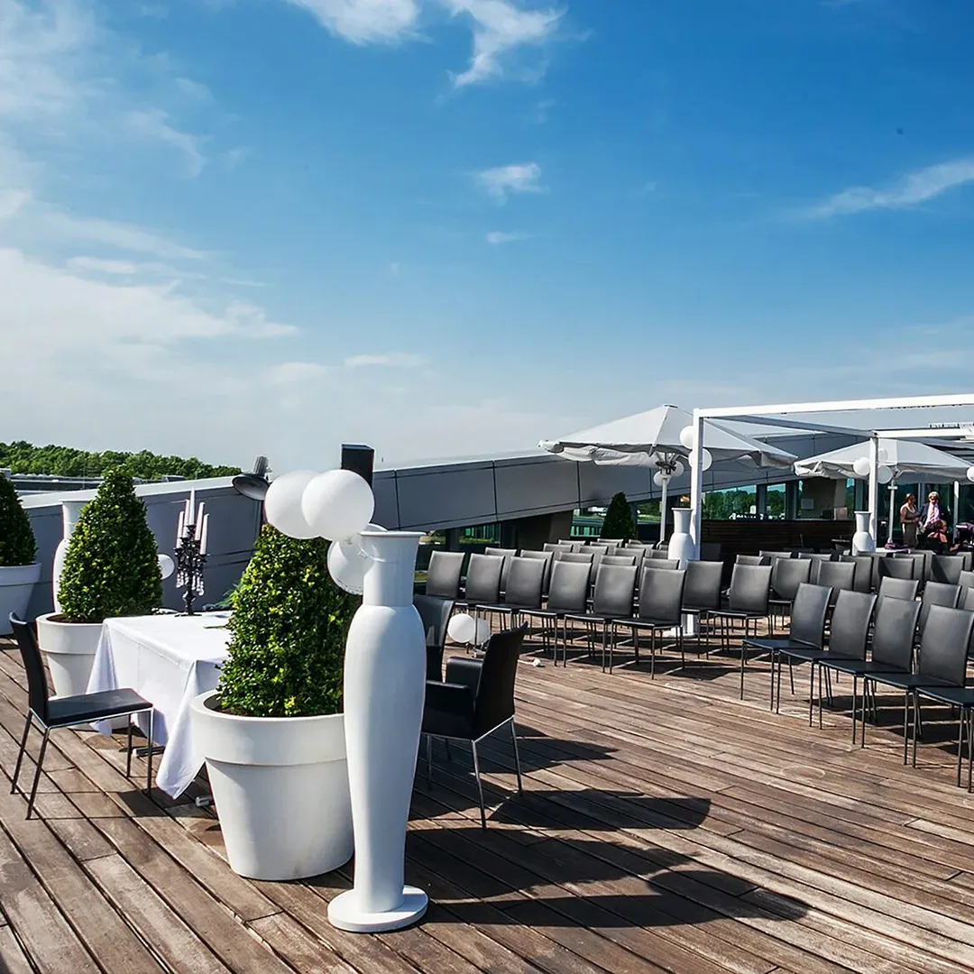 Dachterrasse des Hotel Kameha Grand in Bonn, mit modernen Stühlen, weißen Tischen und dekorativen Pflanzen. Die Sitzgelegenheiten sind für eine Veranstaltung angeordnet, unter einem strahlend blauen Himmel.