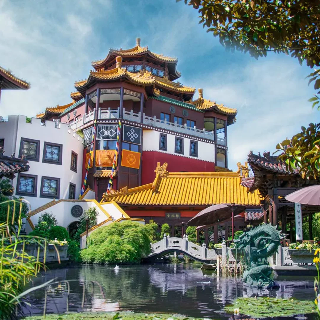 Farbige Außenansicht eines chinesisch inspirierten Gebäudes im Phantasialand in Brühl, Deutschland, mit traditionellen Pagodendächern, leuchtend gelben und roten Akzenten, umgeben von einem Teich und üppiger Vegetation.