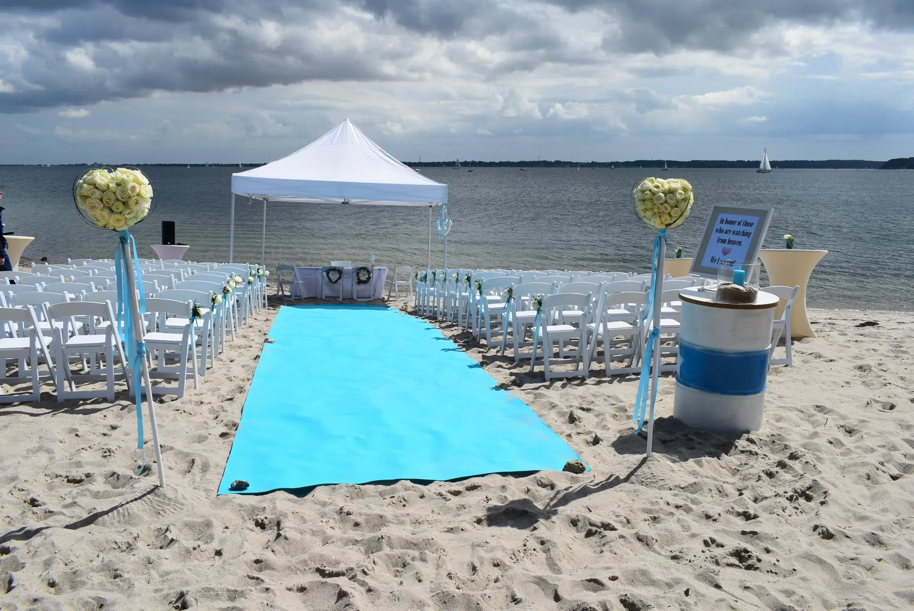 Ein Strandhochzeits-Setup mit einem blauen Teppich, der zu einem weißen Pavillon am Ufer führt. Weiße Stühle und Blumenarrangements säumen den Weg, während im Hintergrund das Meer zu sehen ist.