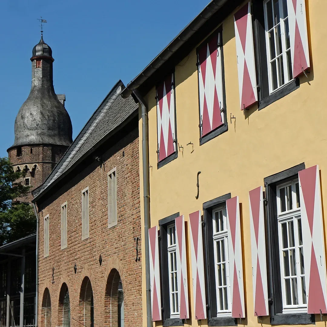 Gelbes Gebäude mit rot-weißen Fensterläden des Kreismuseums in Zons, neben einem mittelalterlichen Turm unter einem klaren, blauen Himmel.