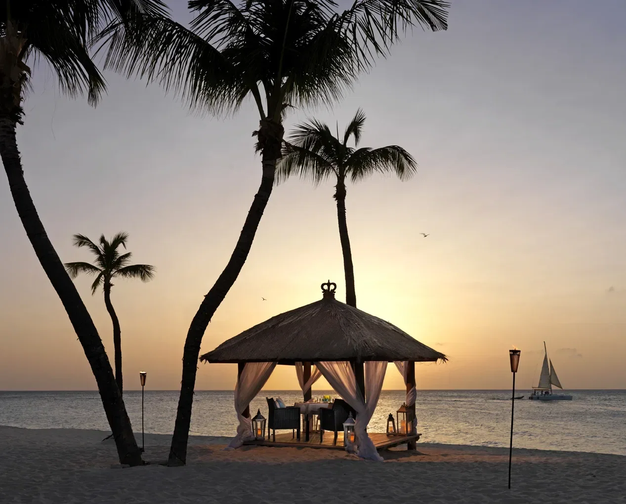 Ein romantischer Pavillon mit Strohdach steht am Strand, beleuchtet von Fackeln, während die Sonne untergeht. Im Inneren sind ein Tisch und Stühle für ein Abendessen mit Blick auf das ruhige Meer arrangiert. Palmen rahmen die Szene ein, und ein Segelboot ist in der Ferne zu sehen.