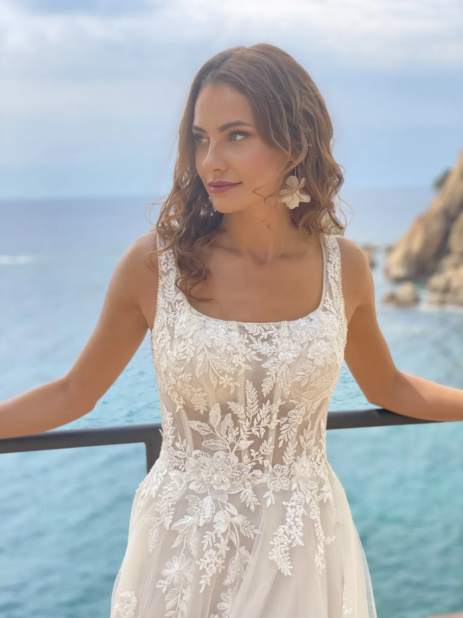 Eine Braut steht vor einem Meerespanorama und trägt ein romantisches Spitzenkleid mit floralen Details. Der fließende Stoff und das zarte Blumenmuster betonen ihre natürliche Schönheit, während das blaue Meer und die Felsen im Hintergrund eine idyllische Kulisse bilden.