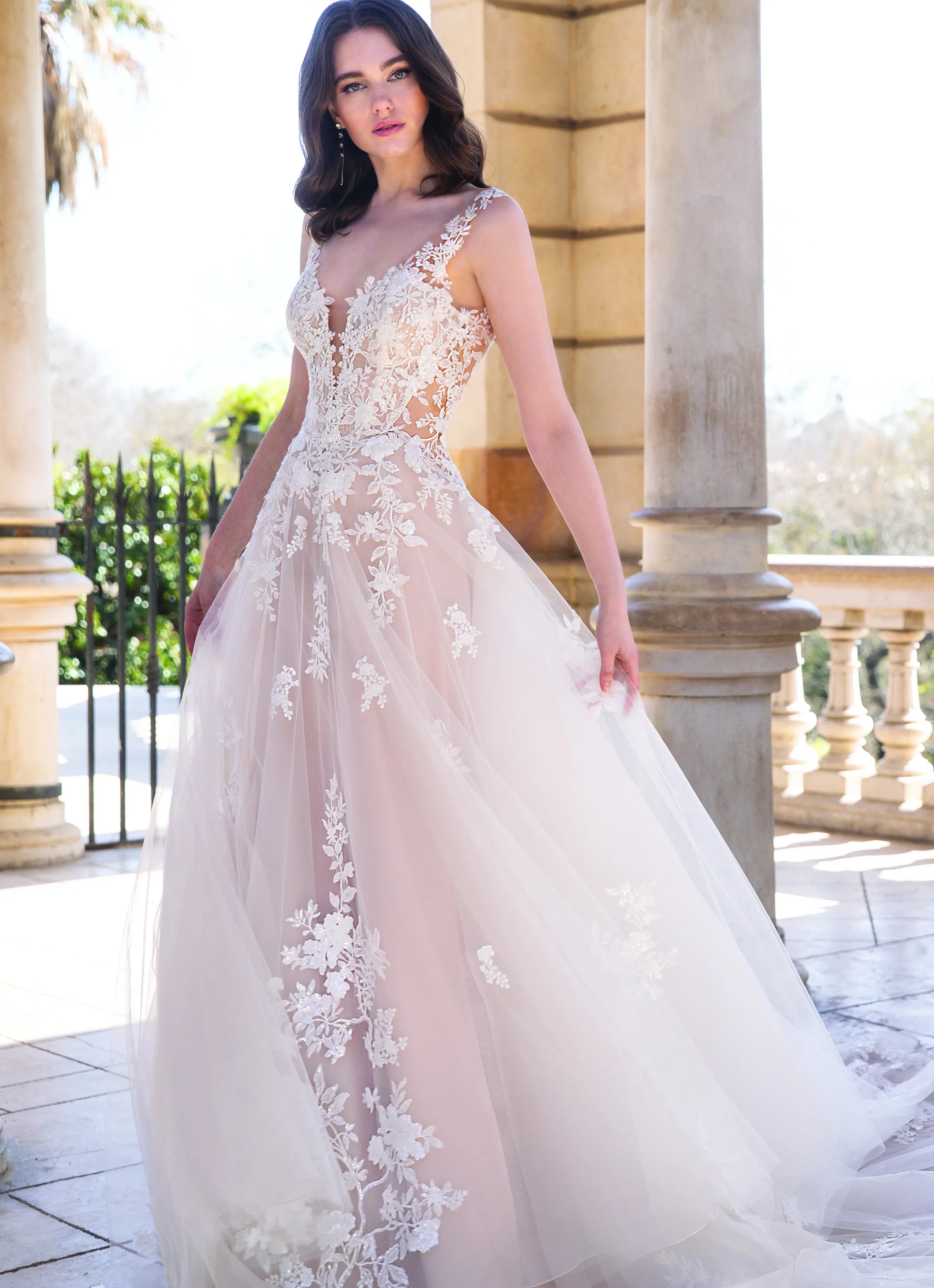 Eine Braut posiert in einem eleganten Hochzeitskleid mit transparenten Spitzenapplikationen auf einem weitläufigen Balkon. Das Kleid mit floralen Verzierungen und einem tiefen V-Ausschnitt fließt anmutig in einem A-Linien-Schnitt, während die klassische Architektur im Hintergrund den edlen Look betont.