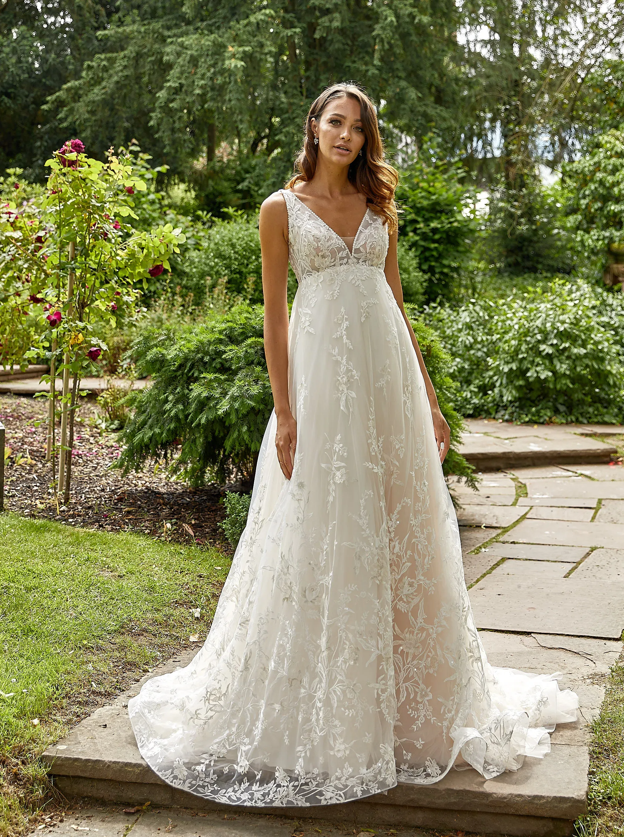 Eine Braut in einem A-Linien-Hochzeitskleid mit V-Ausschnitt und zarter floraler Spitze steht in einem üppigen Garten. Der lange, transparente Rock des Kleides verleiht ihr einen romantischen und eleganten Look, während die umgebende Natur eine friedliche Atmosphäre schafft.