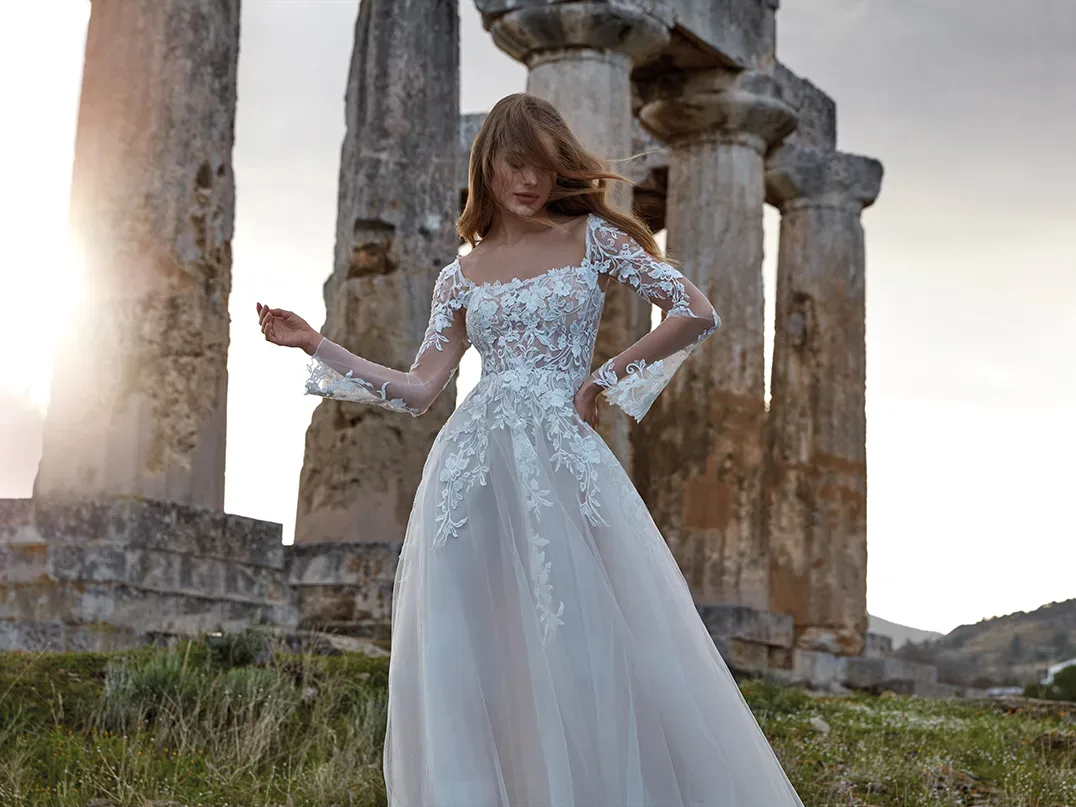 Eine Braut in einem kunstvoll bestickten Kleid mit langen, transparenten Ärmeln steht vor antiken Säulenruinen. Der Wind bewegt leicht ihren Rock, während das Kleid mit floralen Applikationen und einem Carmen-Ausschnitt perfekt zum historischen Ambiente passt.
