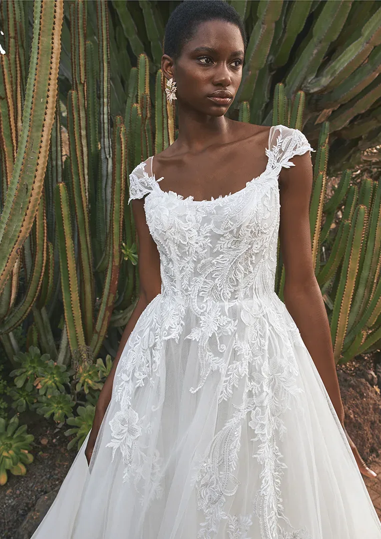 Eine Braut in einem eleganten Spitzenkleid mit transparenten Trägern und floralen Stickereien steht vor großen Kakteen. Das Kleid betont ihre schlichte Eleganz, während die natürliche Kulisse mit den Kakteen einen exotischen Kontrast schafft.
