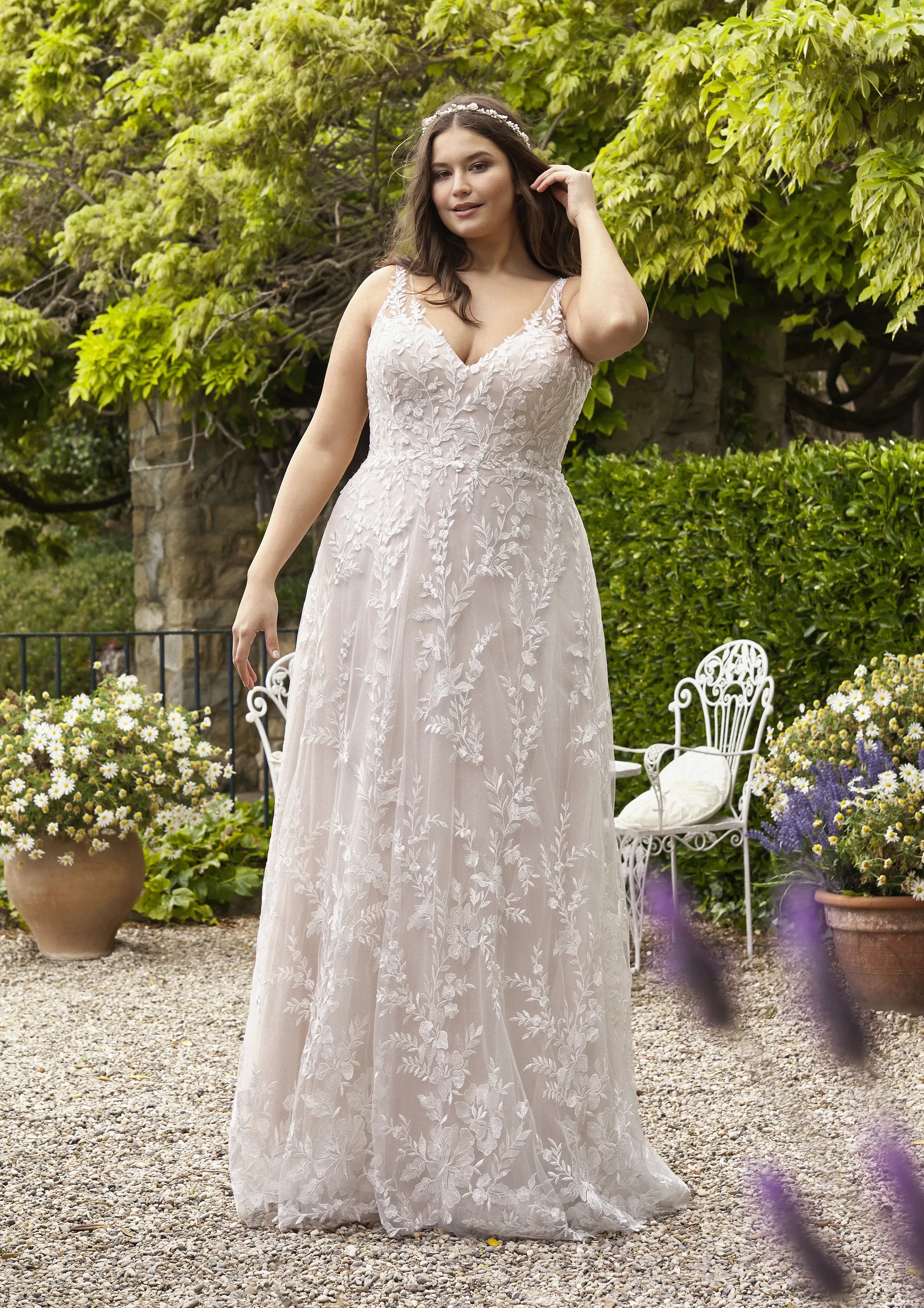 Eine Braut in einem romantischen A-Linien-Kleid mit tiefem Ausschnitt und floraler Spitze steht in einem Garten, umgeben von blühenden Pflanzen. Der fließende Tüllrock und die elegante Schleppe verleihen ihr einen femininen und natürlichen Look.