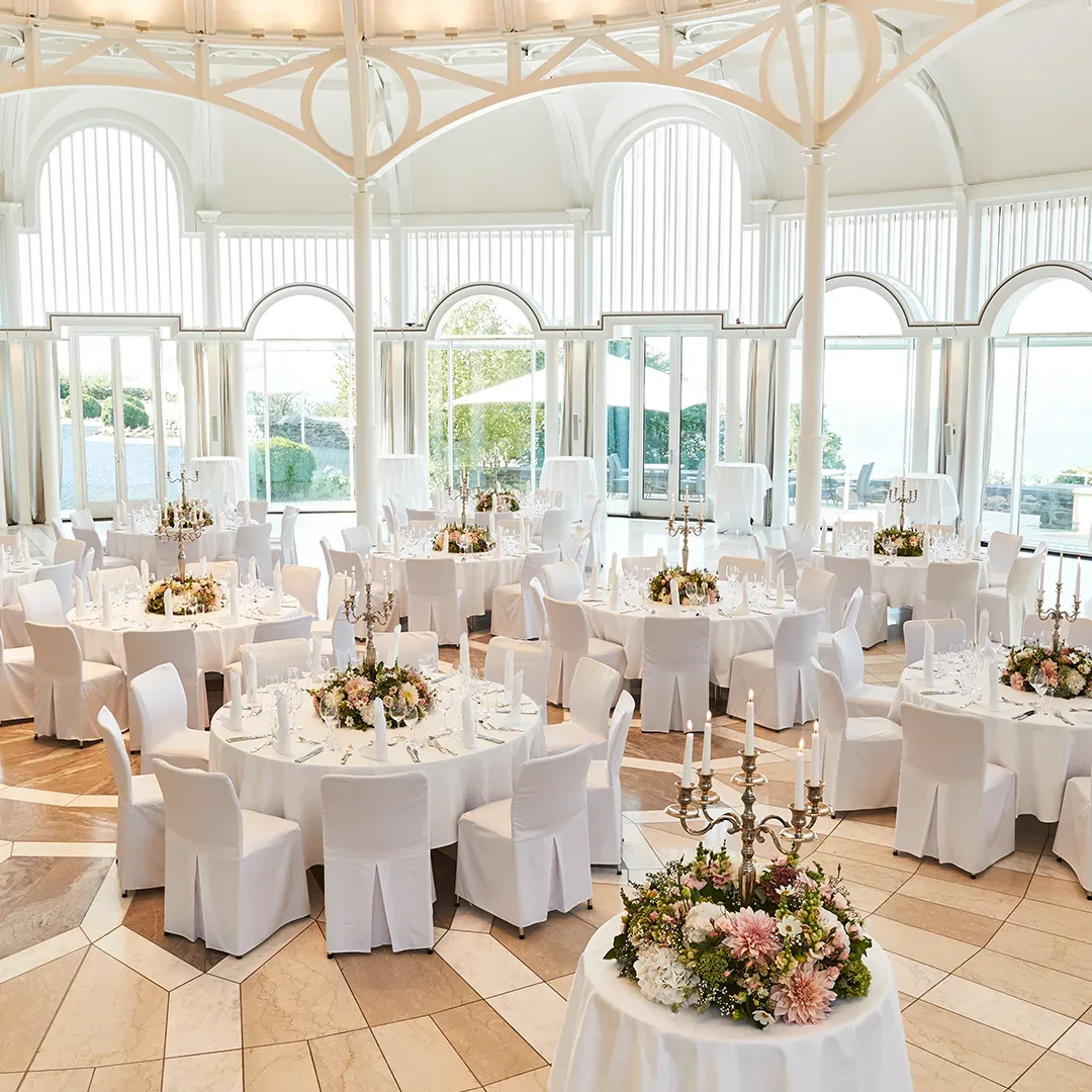 Innenansicht des Festsaals im Grand Hotel Petersberg, Königswinter, mit eleganten runden Tischen, die für eine Hochzeit gedeckt sind, umgeben von großen Fenstern, die Tageslicht in den Raum lassen.