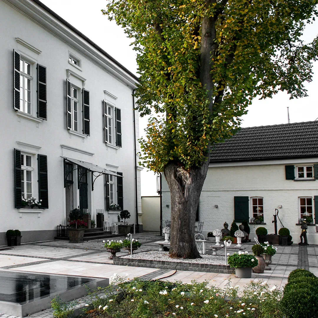 Blick auf den Innenhof von La Remise in Königswinter, einem eleganten weißen Gebäude mit schwarzen Fensterläden und einem zentralen Baum, umgeben von gepflegten Pflanzen und einer Terrasse.