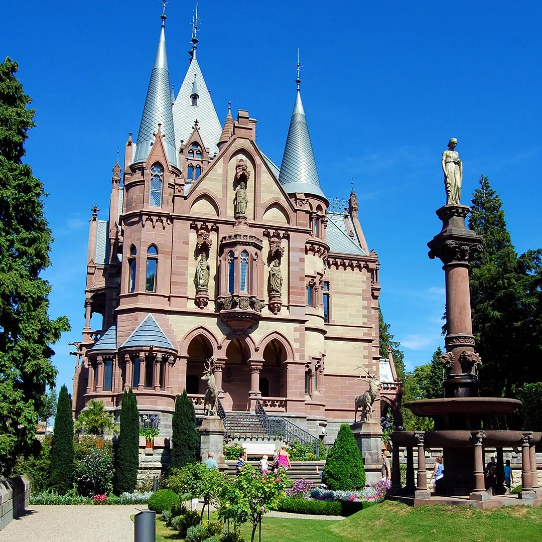 Frontansicht von Schloss Drachenburg in Königswinter, einem prachtvollen neugotischen Schloss mit spitzen Türmen und kunstvollen Steinfiguren, umgeben von gepflegten Gärten, vor einem strahlend blauen Himmel.