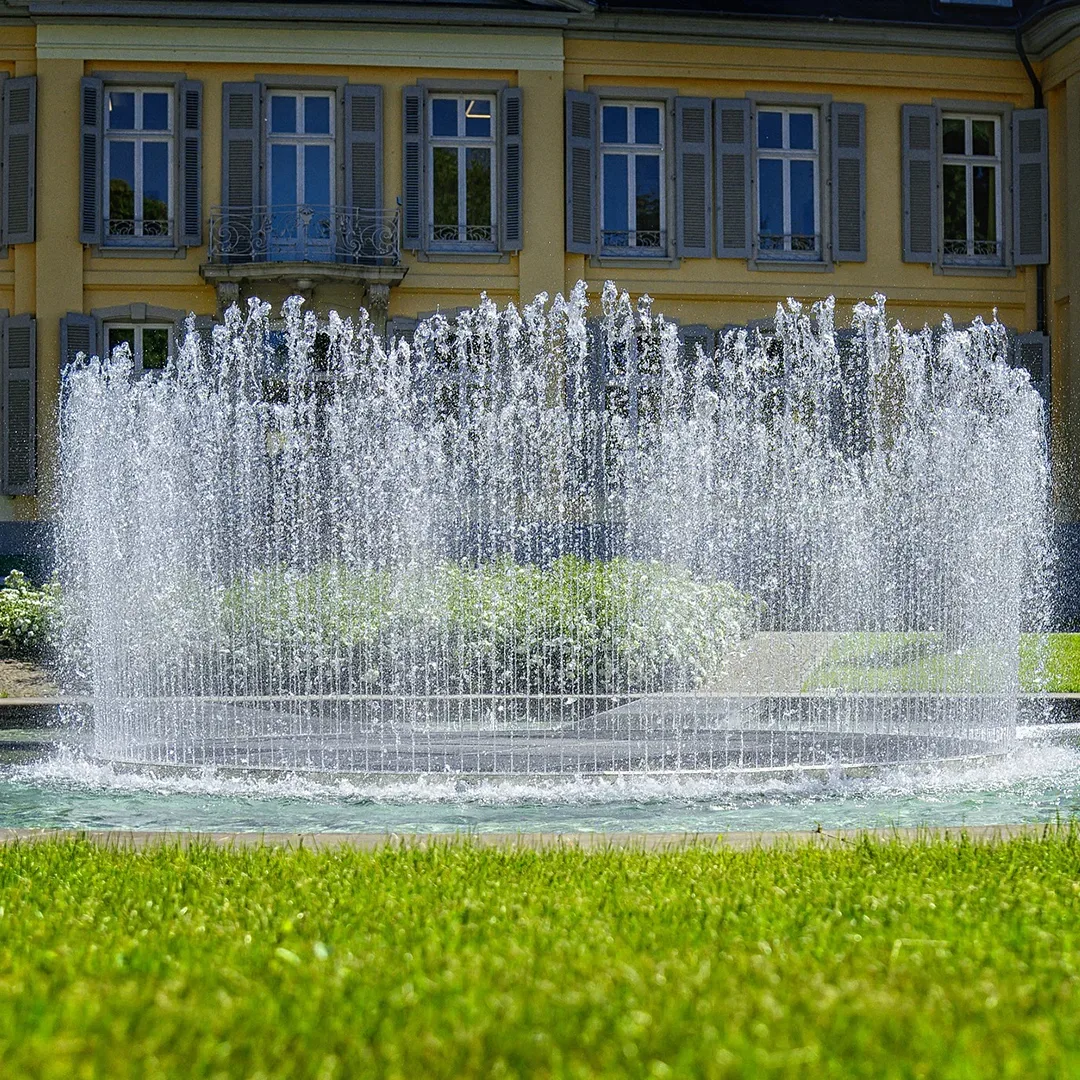 Ein sprudelnder Springbrunnen im Garten des barocken Schloss Morsbroich in Leverkusen, mit der gelben Fassade des Schlosses im Hintergrund.