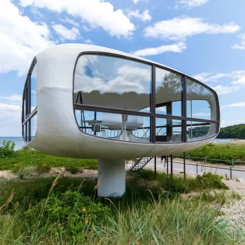 Futuristisches Gebäude in Form eines Pilzes mit großen Glasfenstern, das auf einer Säule am Strand von Binz steht, umgeben von Dünen und blauem Himmel.