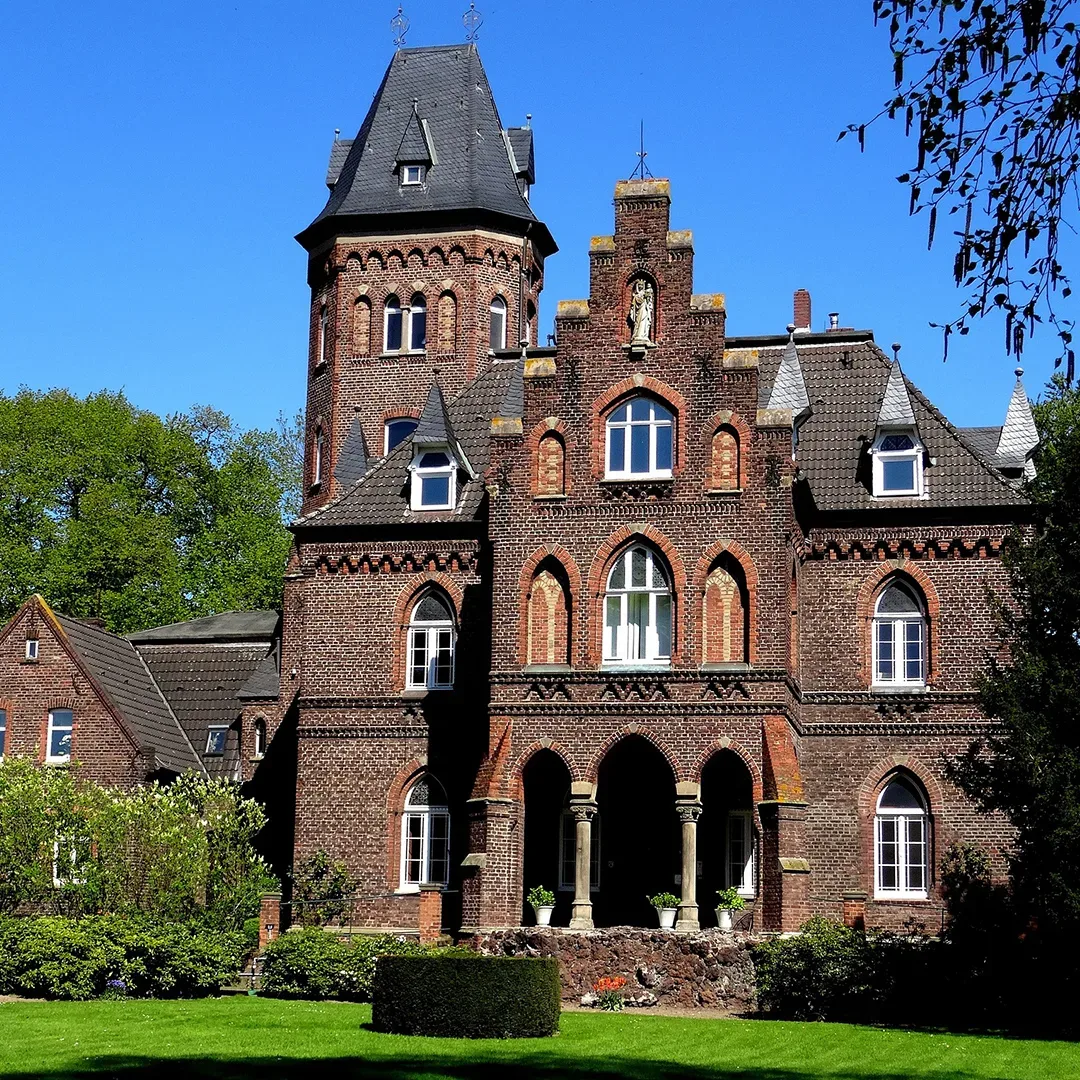 Die beeindruckende Backsteinfassade der Marienburg in Monheim, mit ihrem hohen Turm und neugotischen Architekturdetails unter einem strahlend blauen Himmel.