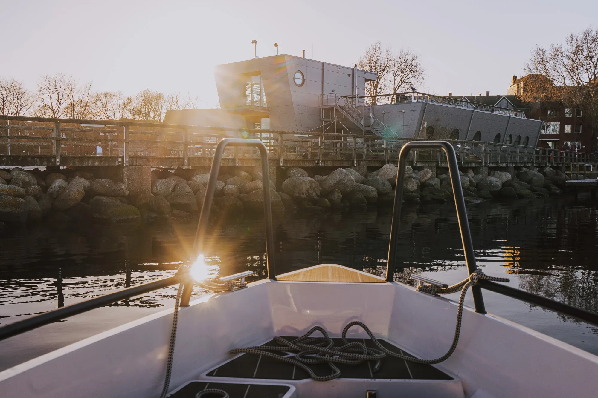 Eine Aufnahme aus der Perspektive des Bootes, das im Hafen liegt. Im Hintergrund erstrahlt die Sonne am Horizont, während ein modernes Gebäude auf einem Steg sichtbar ist.