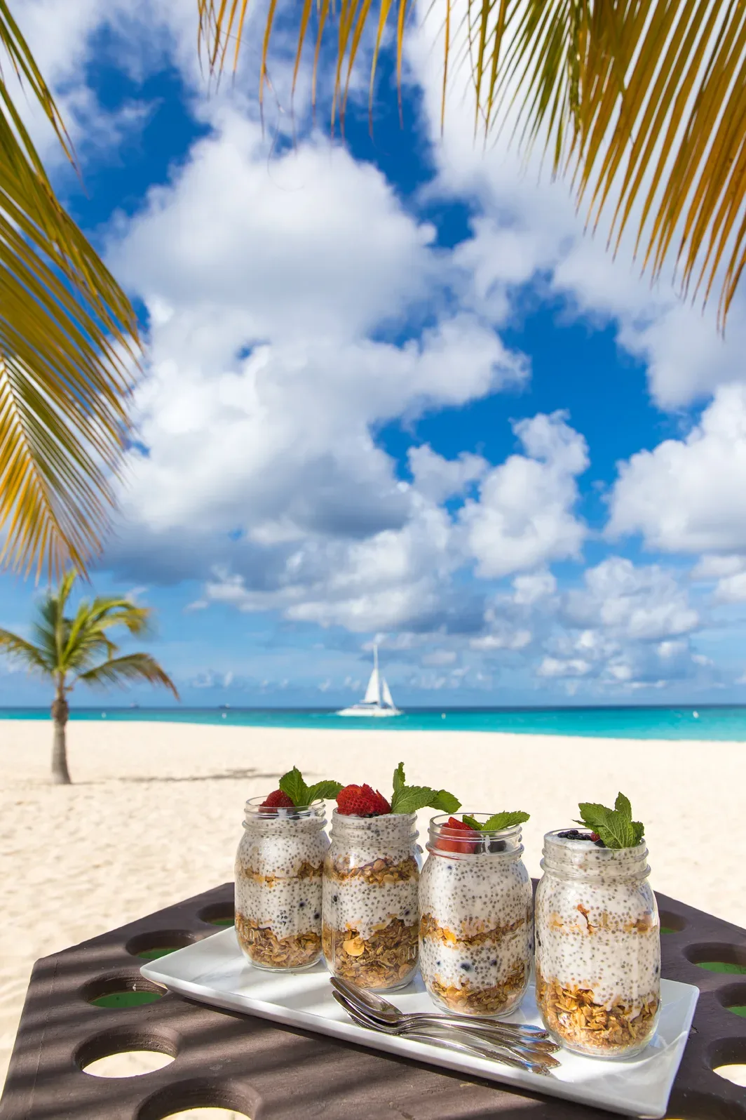 Vier Gläser mit Chia-Pudding, verziert mit Beeren und Minze, stehen auf einem Tablett am Strand unter Palmen. Im Hintergrund sind das türkisfarbene Meer, ein Segelboot und ein strahlend blauer Himmel mit weißen Wolken zu sehen.