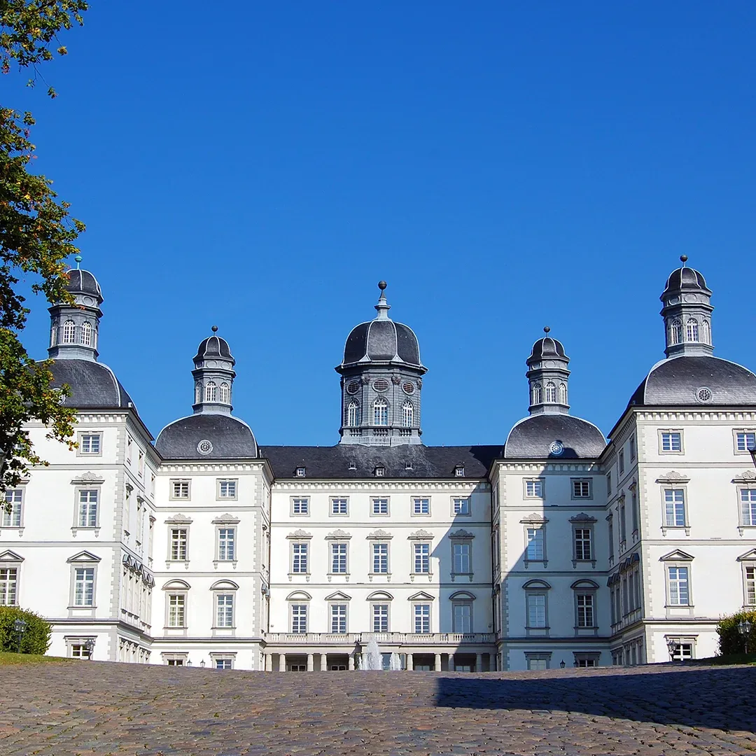 Frontansicht von Schloss Bensberg, einem prächtigen barocken Schloss in Bergisch Gladbach, Deutschland, mit mehreren Kuppeltürmen und einer symmetrischen weißen Fassade unter einem klaren blauen Himmel.