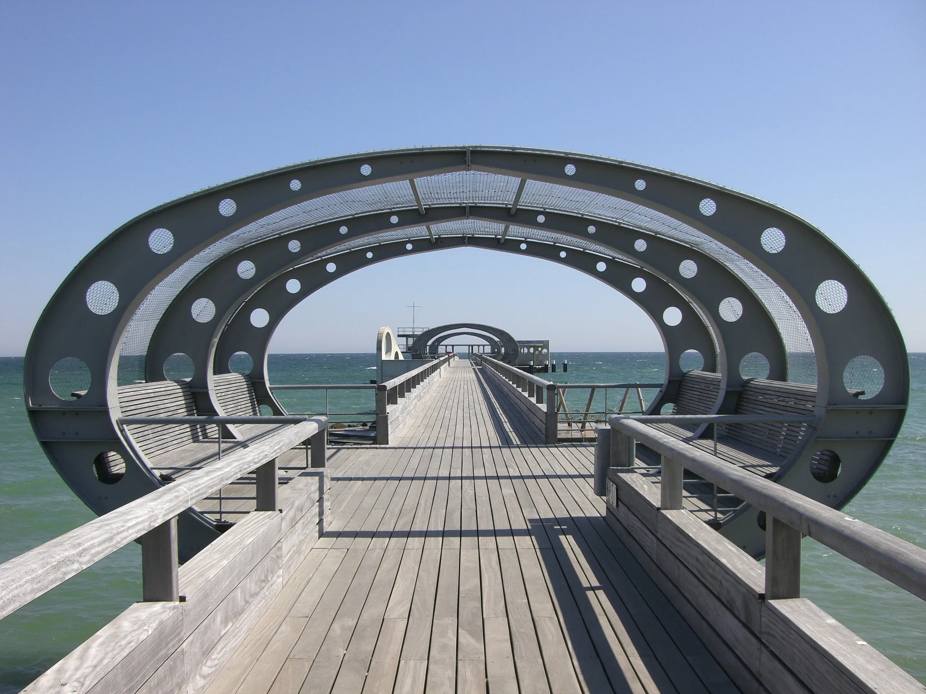 Moderne Seebrücke an der Ostsee in Kellenhusen mit futuristischem, rundem Design. Die Brücke führt weit ins Meer hinaus, umgeben von klarem, blauem Wasser unter strahlendem Himmel.