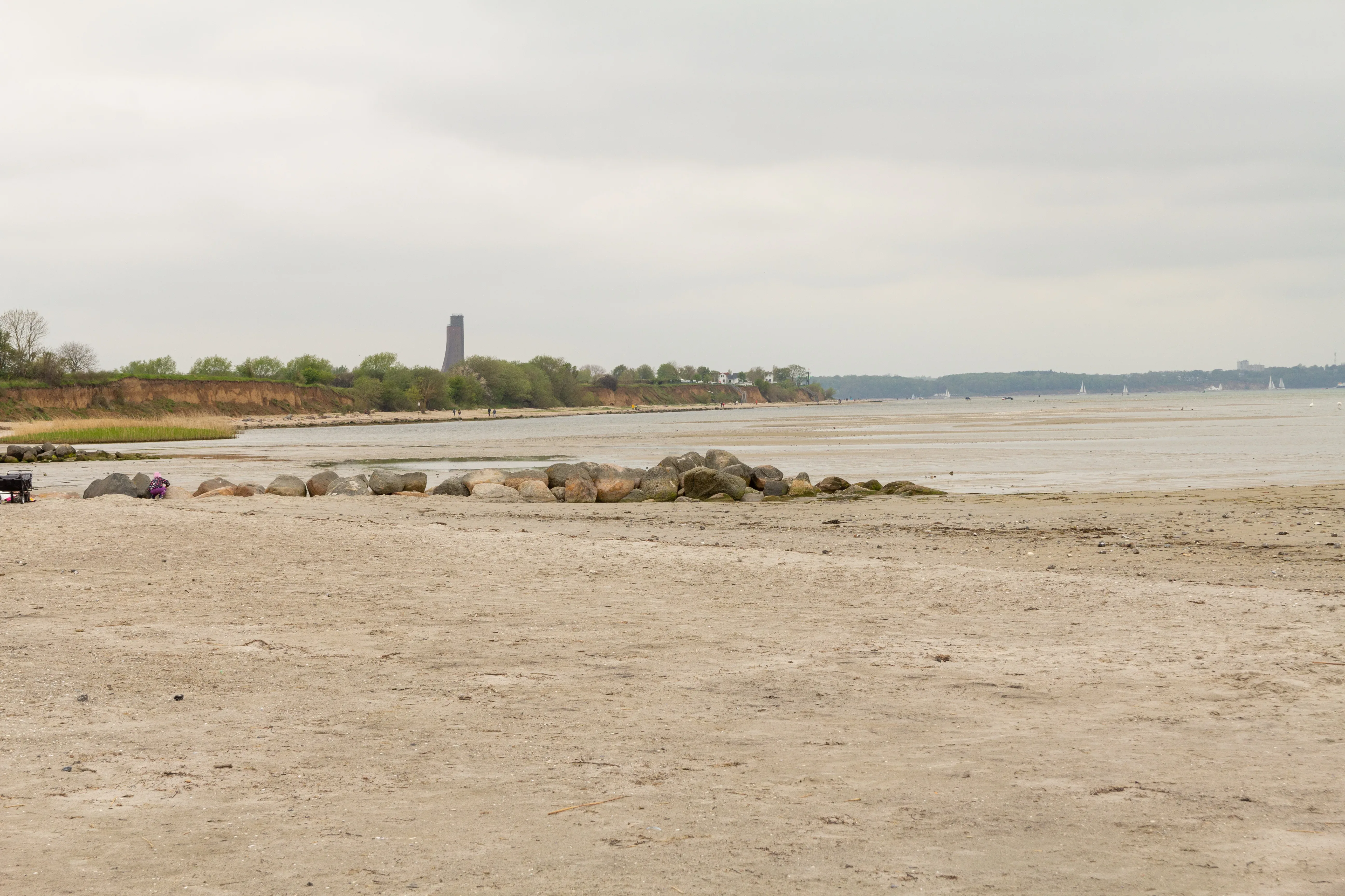Ein weiter Sandstrand mit Felsen im Vordergrund und einer sanften Klippenlinie im Hintergrund. Das Meer erstreckt sich bis zum Horizont unter einem bewölkten Himmel.