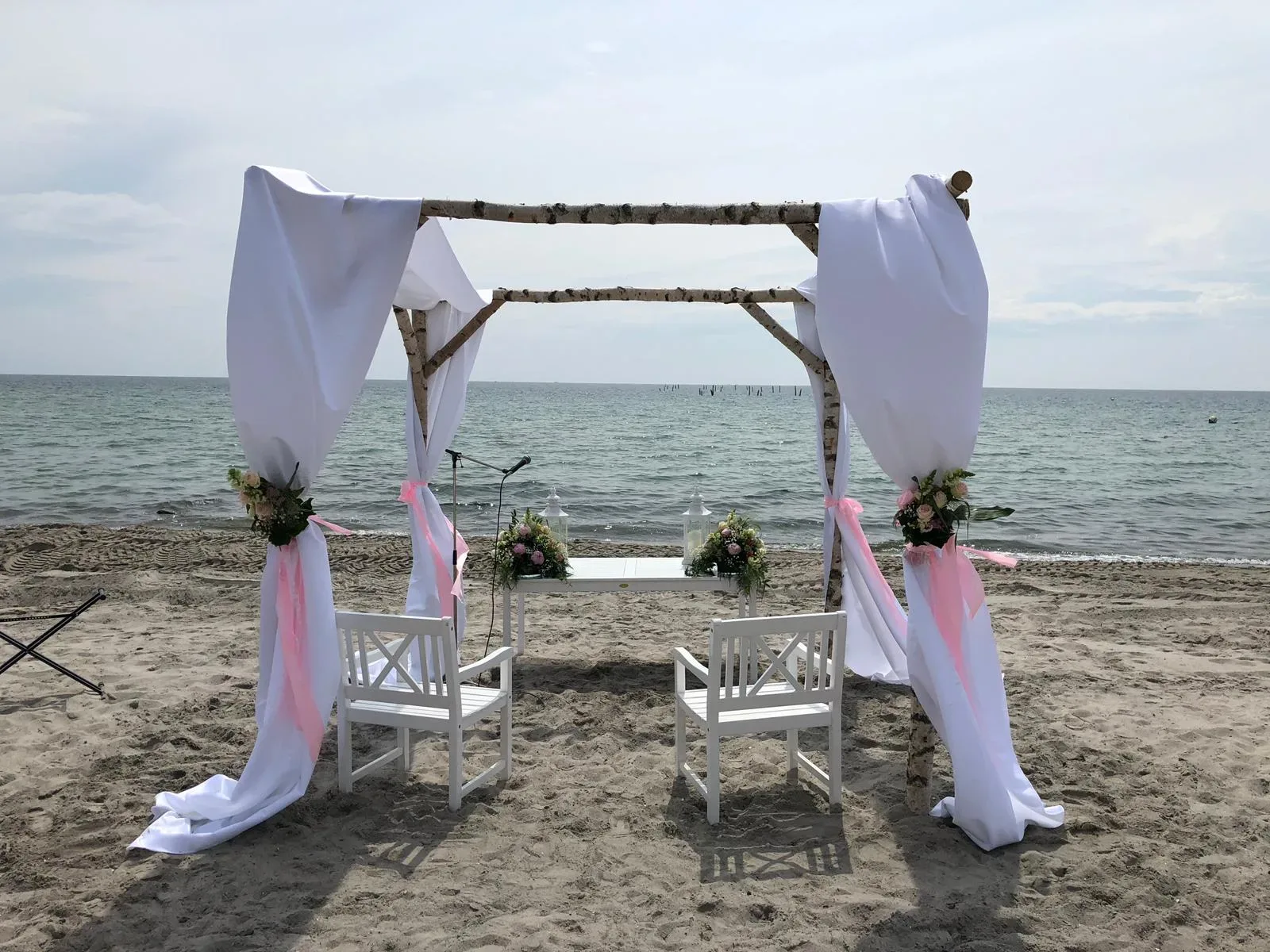 Eine Hochzeitszeremonie am Strand mit einem weißen, geschmückten Pavillon, der mit rosa Bändern und Blumen dekoriert ist. Zwei weiße Stühle stehen bereit, mit Blick auf das Meer.