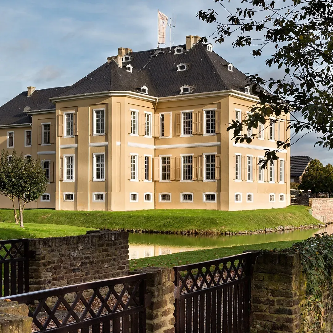 Vorderansicht des Schlosses Miel in Swisttal, einem gelben barocken Herrenhaus mit symmetrischen Flügeln, das von einem Wassergraben und gepflegten Grünflächen umgeben ist.