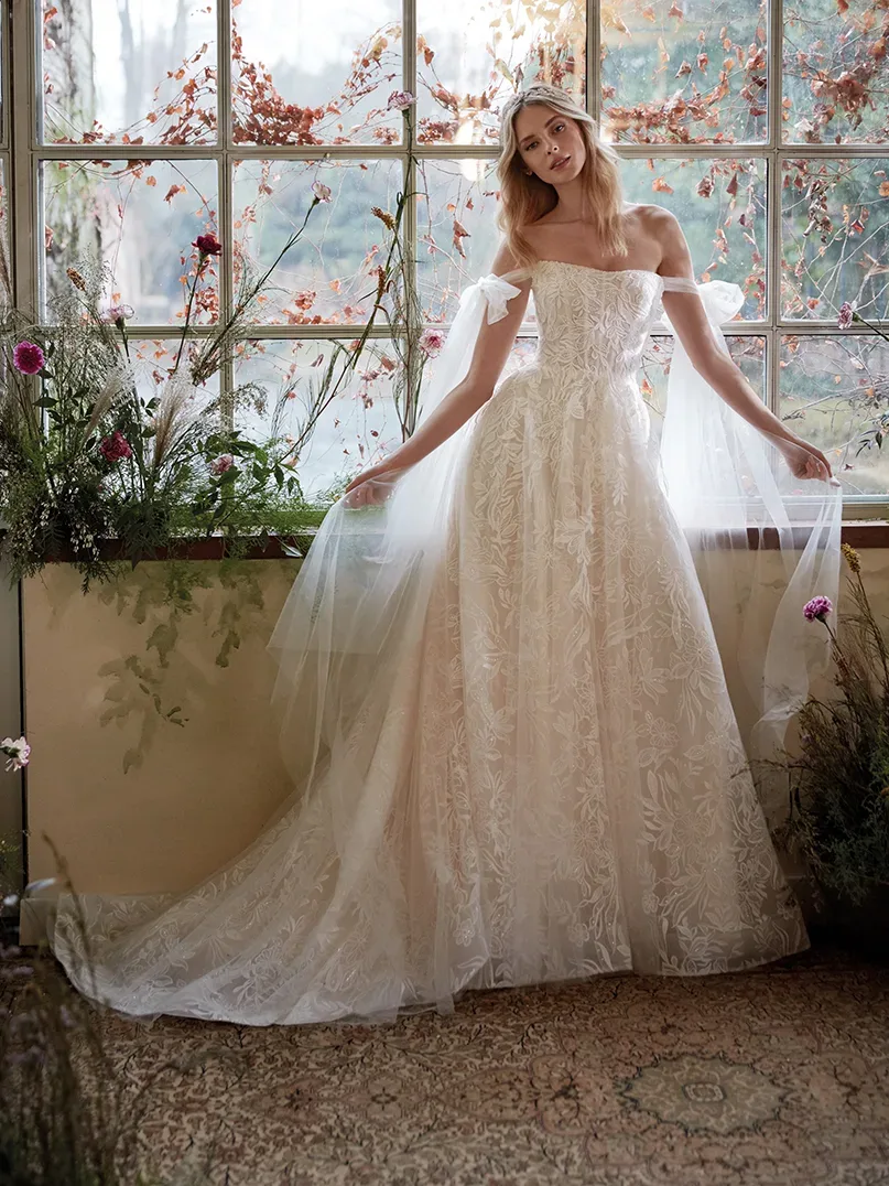Braut in einem märchenhaften Spitzenkleid mit floralen Applikationen und schulterfreiem Design steht vor einem großen Fenster, umgeben von herbstlicher Dekoration. Das Kleid mit Tüllschleier und floralen Details schafft eine verträumte Atmosphäre.