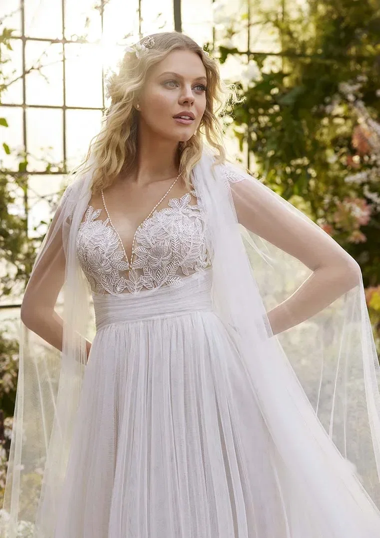 Eine Braut steht in einem lichtdurchfluteten Garten, gekleidet in einem eleganten Hochzeitskleid mit tiefem V-Ausschnitt und floralen Stickereien. Der weiche Tüll und die zarten, durchsichtigen Ärmel verleihen ihr einen romantischen und märchenhaften Look.