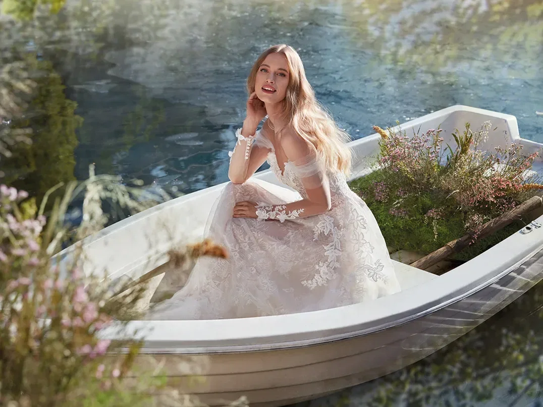 Eine Braut sitzt in einem Ruderboot, umgeben von Blumen und sanften Wasserwellen, gekleidet in ein romantisches Spitzenkleid mit floralen Applikationen und einem schulterfreien Design. Die Szene wirkt märchenhaft und verträumt.