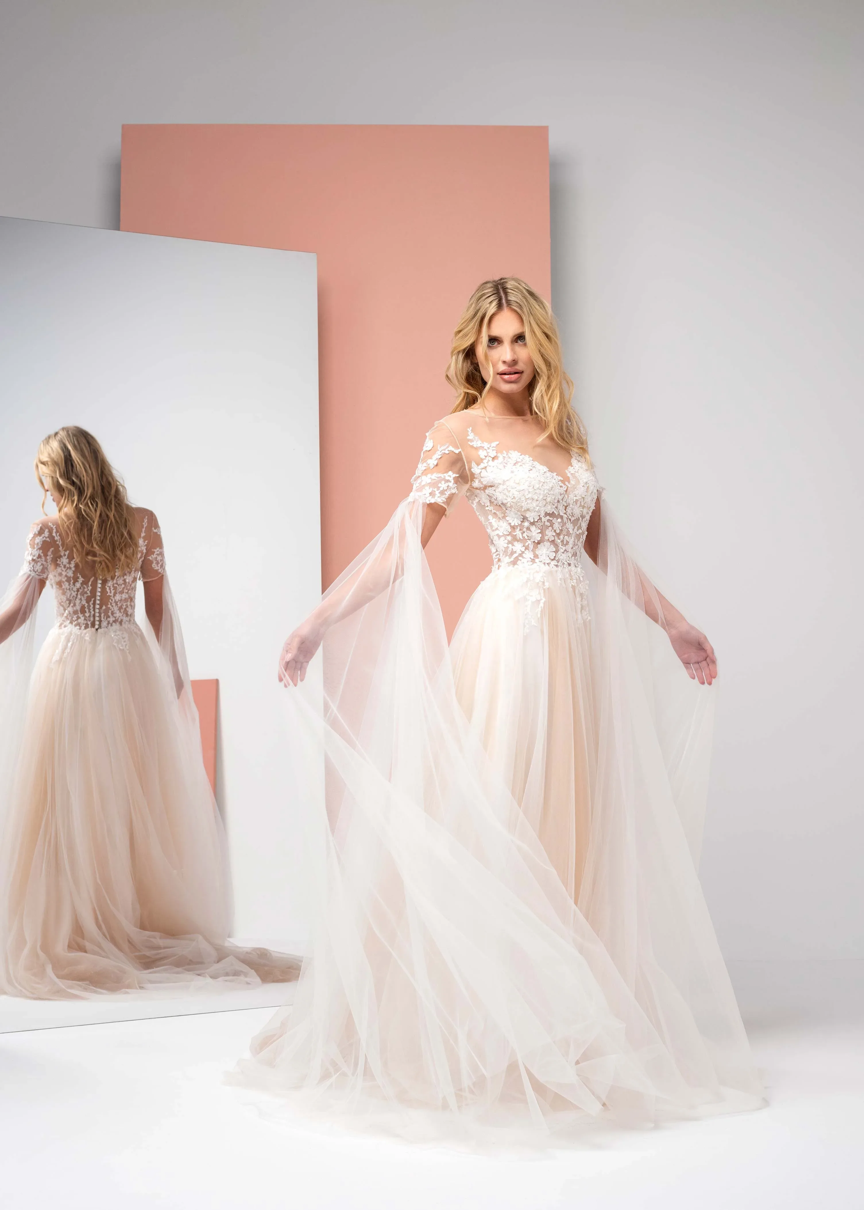Braut in einem modernen Tüllkleid mit langen, transparenten Ärmeln und floralen Applikationen posiert vor einem großen Spiegel. Das helle, minimalistische Design des Raumes und das fließende Kleid sorgen für eine elegante und zeitgemäße Stimmung.