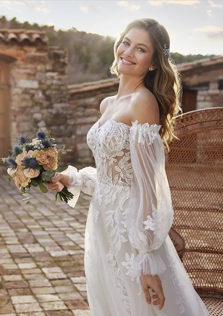 Braut in einem trägerlosen Hochzeitskleid mit floraler Spitze und transparenten Ärmeln, die ein Bouquet aus blauen und beigen Blumen hält.