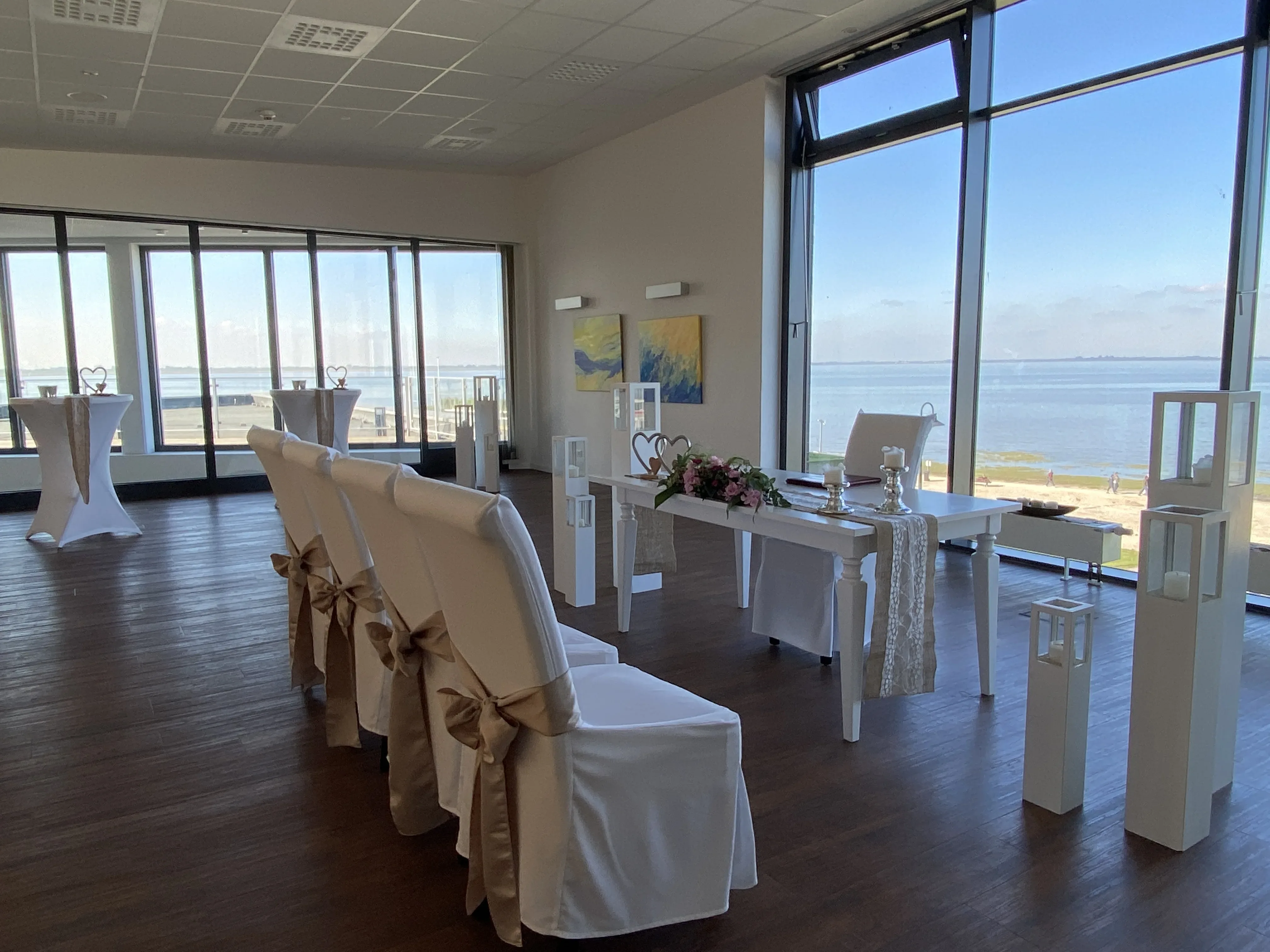 Modernes Trauzimmer mit großen Panoramafenstern, die einen weiten Blick auf das Meer bieten. Weiß dekorierte Stühle und ein festlich geschmückter Tisch stehen im Raum, der mit natürlichen Licht durchflutet wird.