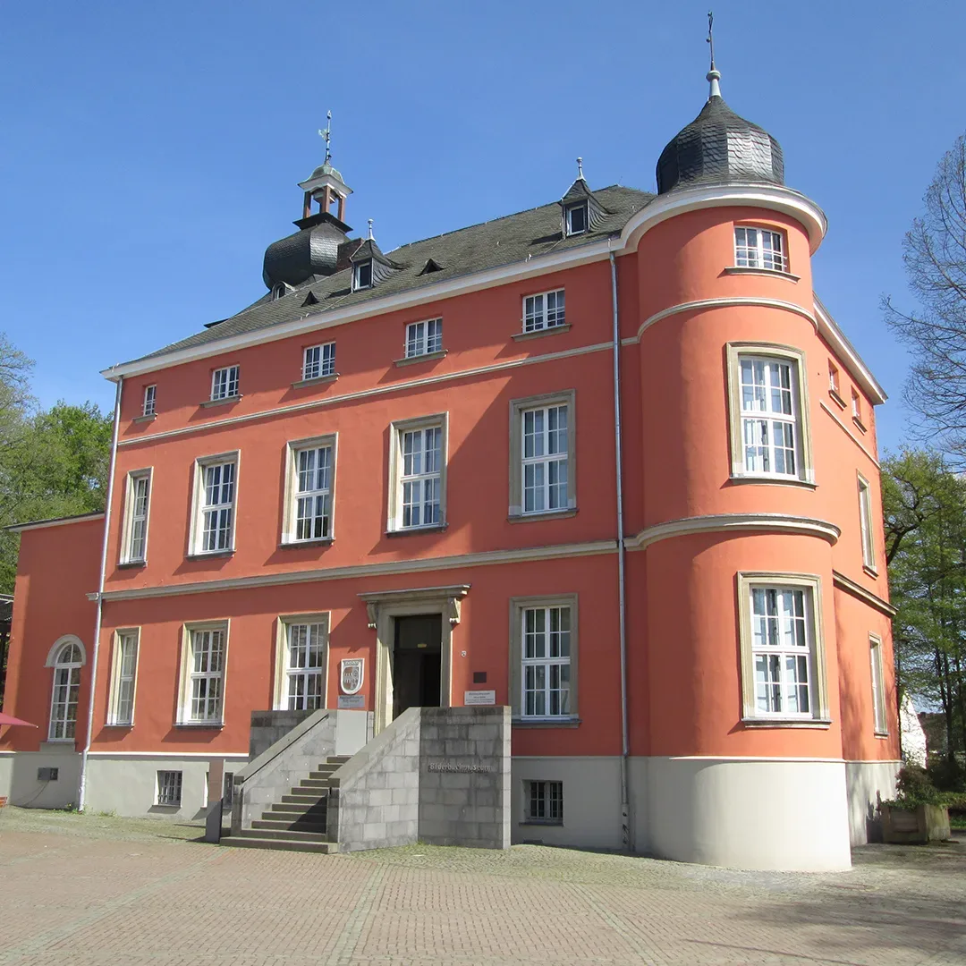 Vorderansicht der Burg Wissern in Troisdorf, einem kleinen roten Backsteinschloss mit rechteckiger Struktur, Ecktürmen und einer Steintreppe, die zum Haupteingang führt, unter einem strahlend blauen Himmel.