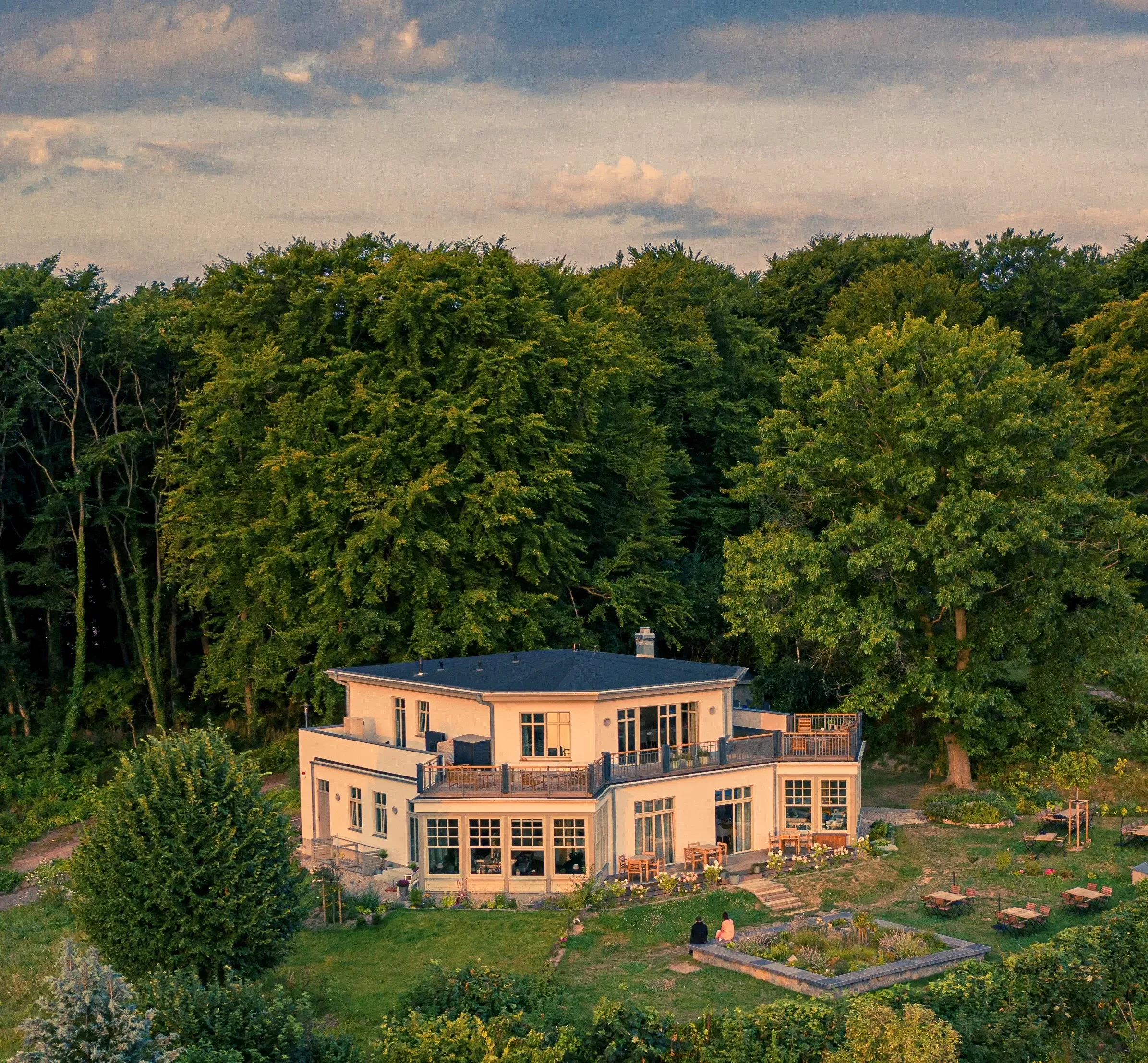 Eine moderne zweistöckige Villa, umgeben von dichtem Wald, aufgenommen während des Sonnenuntergangs. Die Villa hat große Fenster und eine Terrasse, die in die Natur eingebettet ist.