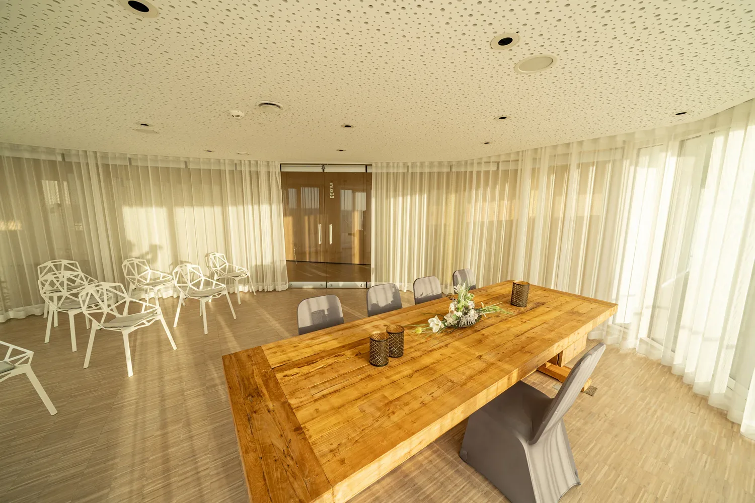 Helles, modernes Trauzimmer mit einem großen Holztisch und Designerstühlen, umgeben von bodenlangen, transparenten Vorhängen, die sanftes Licht in den Raum lassen.