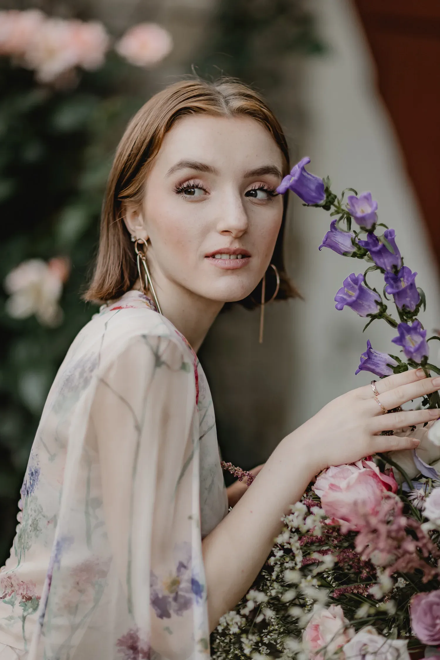 Eine Frau in einem zarten, blumigen Kleid schaut verträumt zur Seite und hält eine violette Blume in der Hand, während sie von weiteren Blumen umgeben ist.