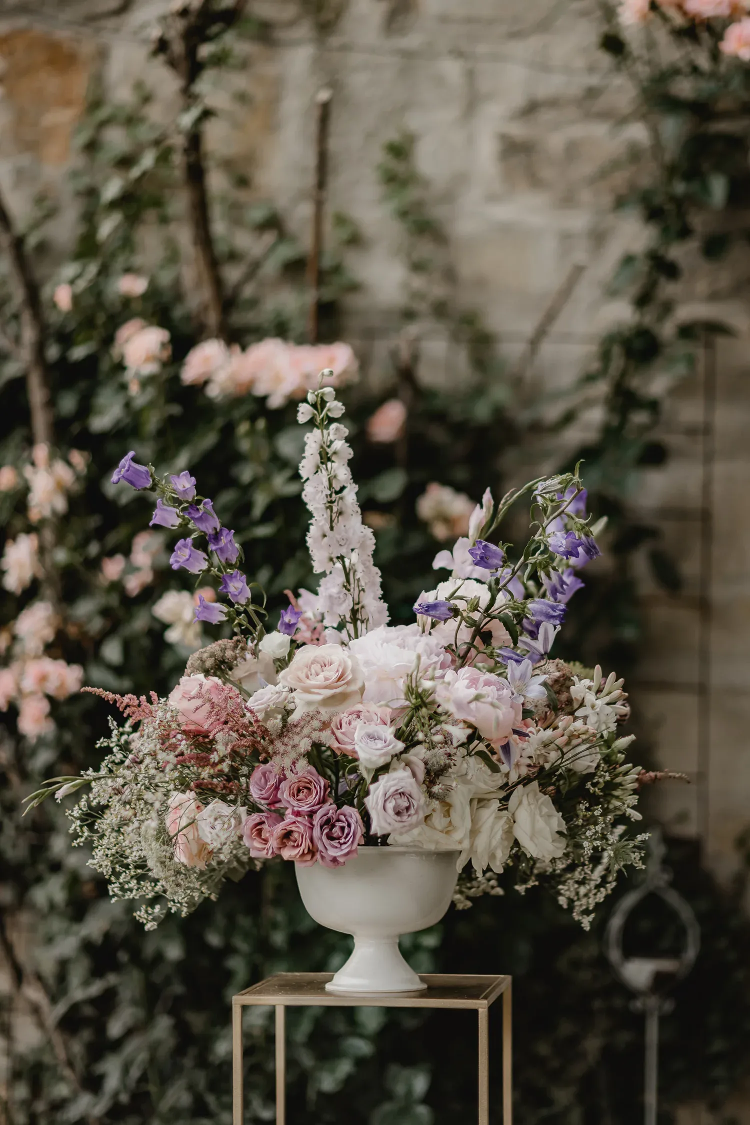 Ein üppiges Blumenarrangement mit Rosen, Glockenblumen und Astern steht in einer weißen Vase vor einer mit Efeu bewachsenen Mauer.