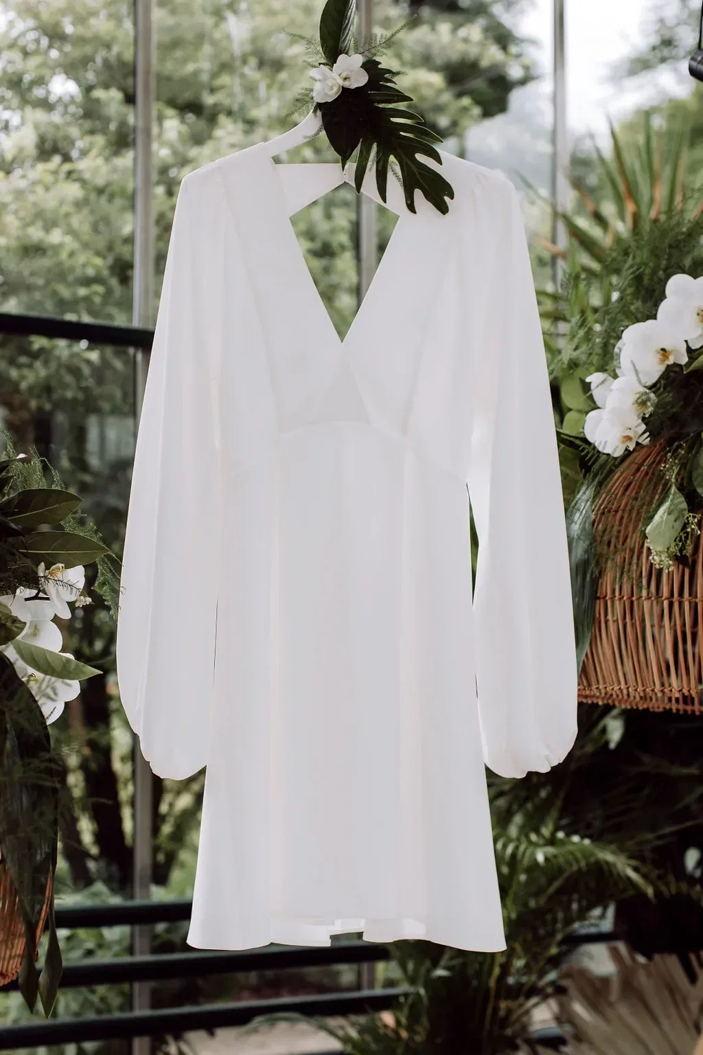 Luftiges, weißes Brautkleid mit V-Ausschnitt, das in einem tropischen Gewächshaus mit Orchideen und Monstera-Blättern hängt.