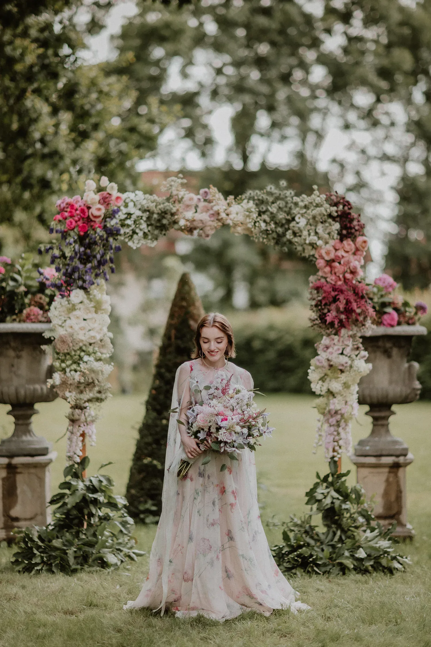 Braut in einem romantischen, blumigen Kleid steht unter einem Blumenbogen im Freien und hält einen großen Blumenstrauß, umgeben von üppiger grüner Natur.