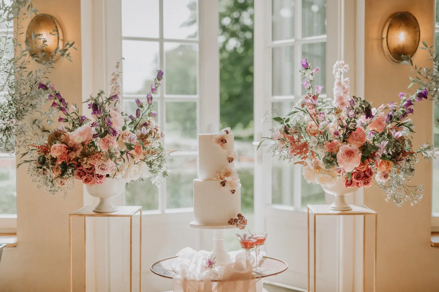 Ein festlich dekorierter Hochzeitstisch mit einer zweistöckigen weißen Torte und zwei Blumenarrangements aus rosa und violetten Blüten, platziert vor bodentiefen Fenstern.