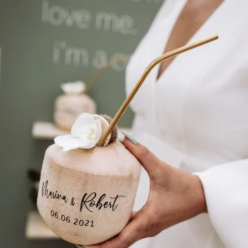 Nahaufnahme einer personalisierten Kokosnuss mit Strohhalm und Blume, die von einer Braut bei einer tropischen Bali-Hochzeit gehalten wird.