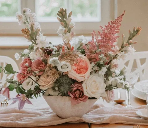 Elegantes Blumenarrangement in Pastelltönen mit Rosen, Astilben und Eukalyptus auf einem festlich gedeckten Tisch, umgeben von Kerzen und Kristallgläsern.