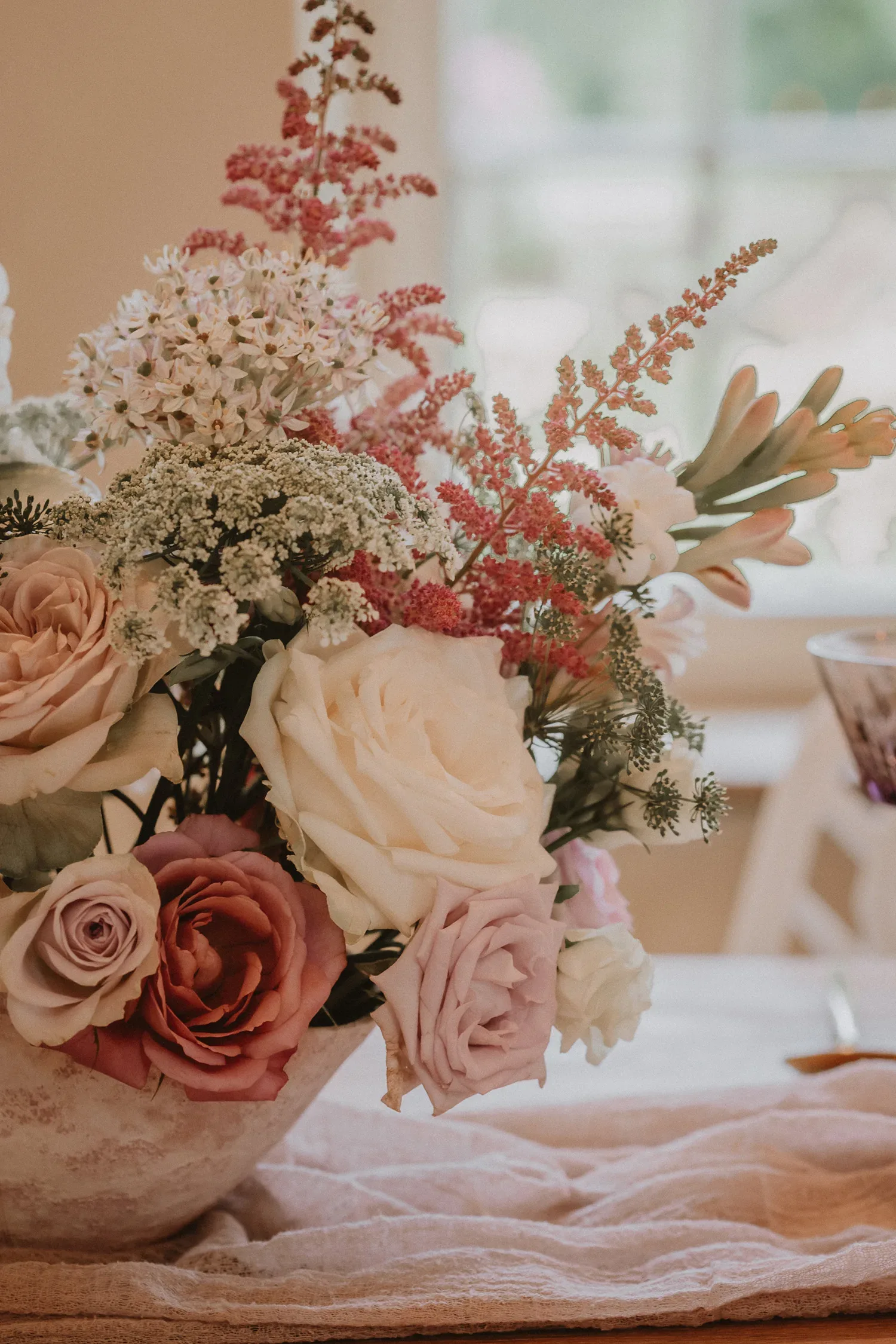 Detailaufnahme eines Blumenarrangements mit großen, pastellfarbenen Rosen, Astilben und weißen Wildblumen in einer Schale auf einem mit Stoff bedeckten Tisch.