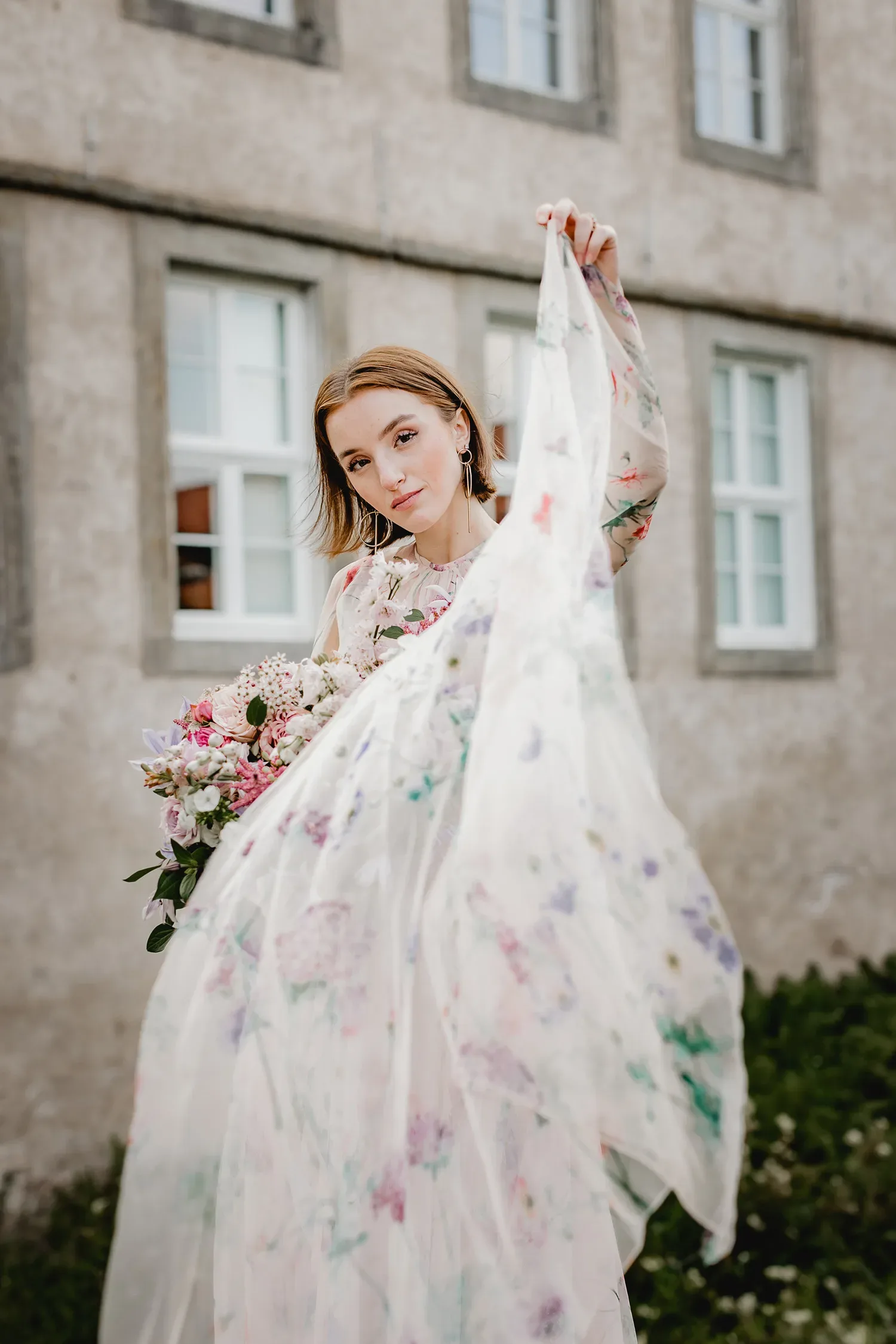 Eine junge Frau in einem geblümten Kleid wirft einen verträumten Blick auf die Kamera, während sie das Kleid elegant zur Seite hält und einen Blumenstrauß vor einem historischen Gebäude trägt.